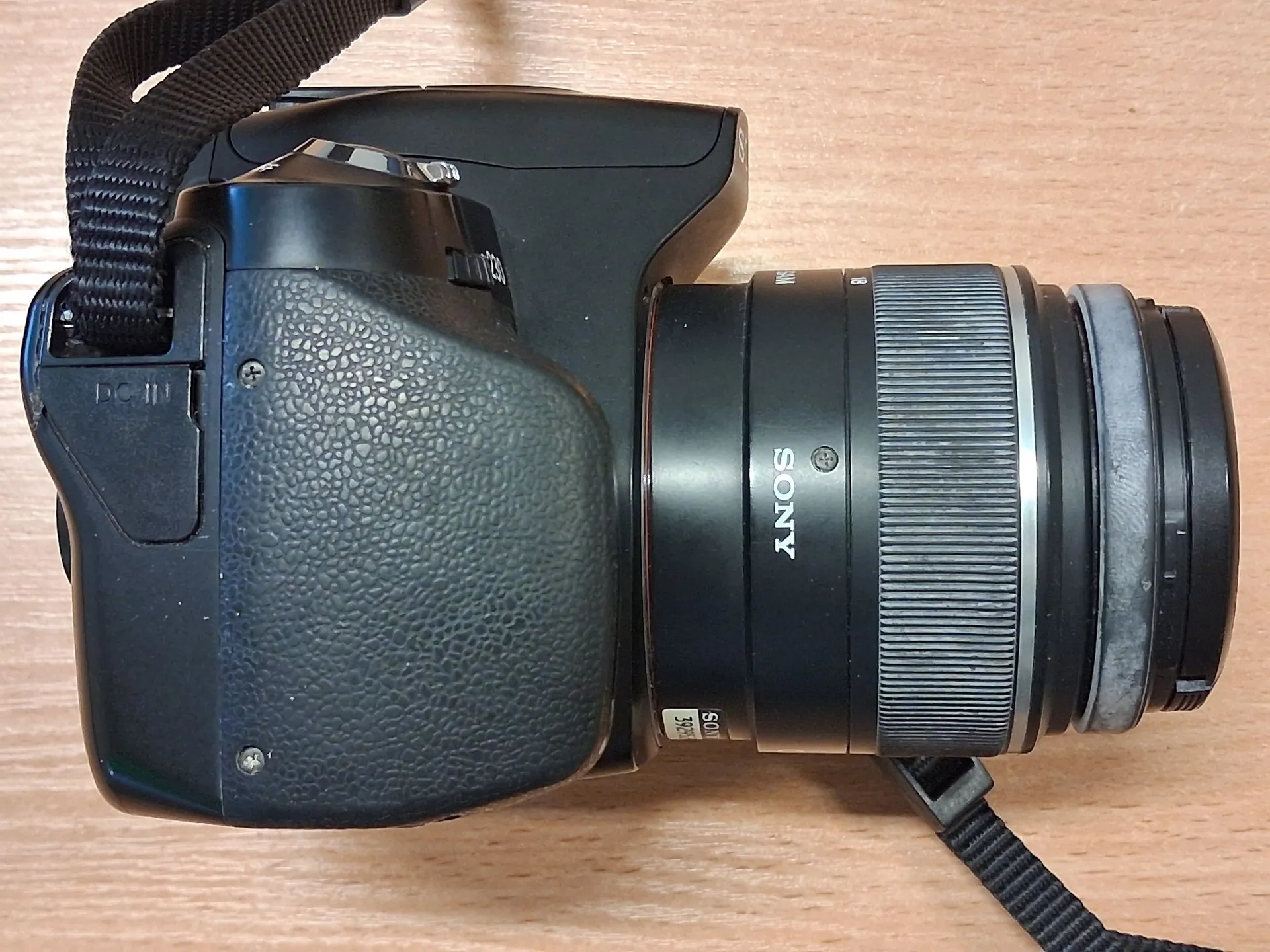 Czarny aparat fotograficzny marki Sony z obiektywem, leżący na drewnianej powierzchni. Na obiektywie widoczny jest napis 'SONY'. Aparat ma przymocowany czarny pasek transportowy.