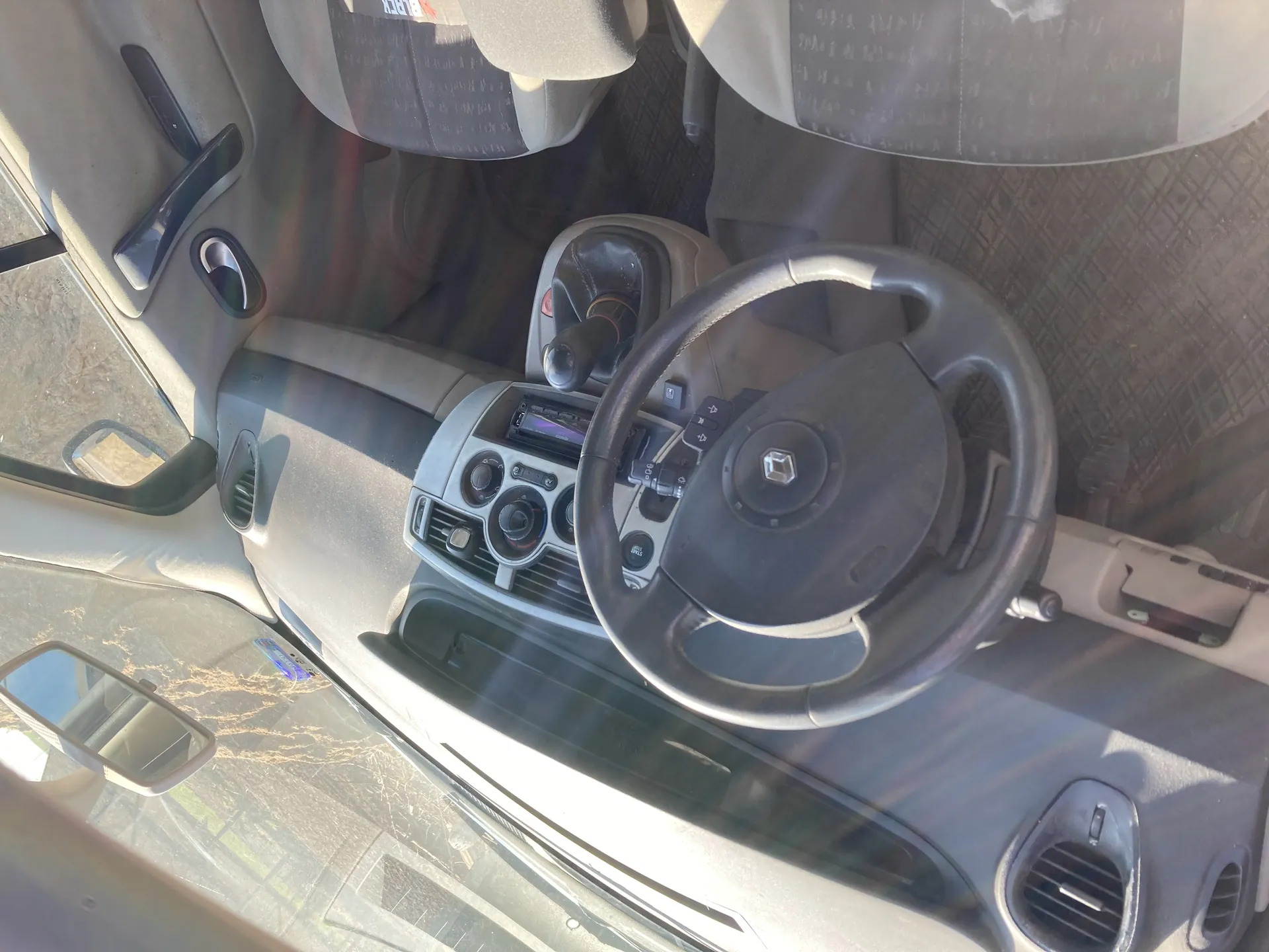 Wnętrze samochodu z kierownicą z logo w centrum, deską rozdzielczą z przyciskami i wyświetlaczem, dźwignią zmiany biegów, siedzeniami z wzorzystymi pokrowcami, uchwytem drzwiowym i lusterkiem bocznym. Widok przez przednią szybę na drzewa.