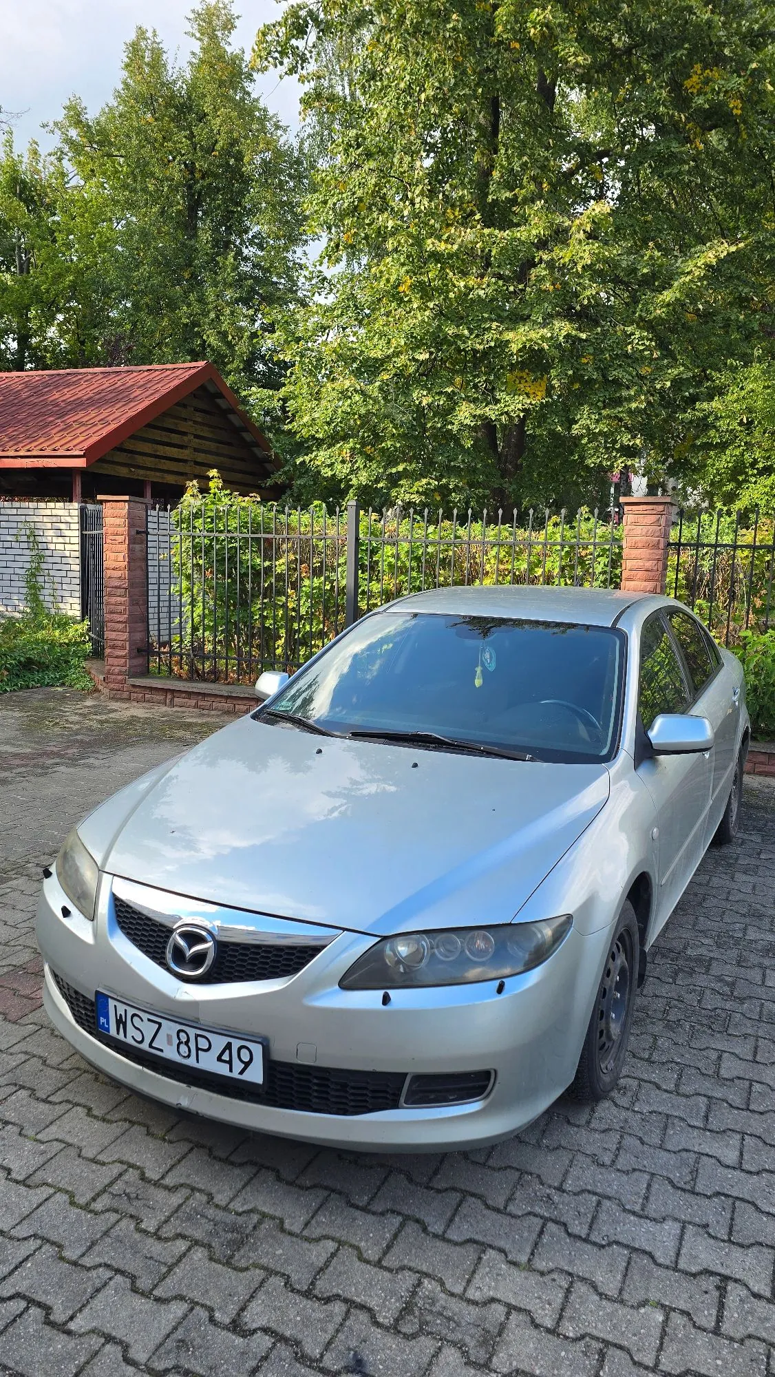 Srebrny samochód marki Mazda zaparkowany na kostce brukowej przed ogrodzeniem i zielonymi drzewami. Na przodzie auta widoczna jest tablica rejestracyjna o numerze WSZ 8P49.