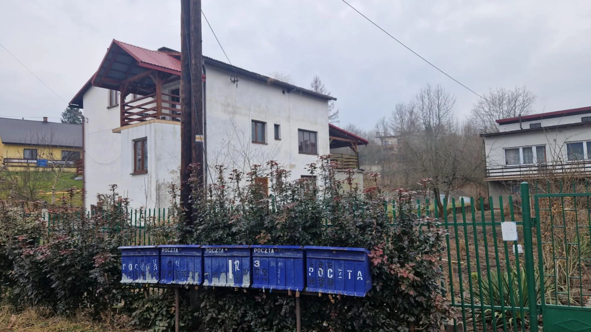 Biały dom z czerwonym dachem i drewnianym daszkiem na balkonie, przed nim zielone ogrodzenie i krzewy. W pierwszym planie niebieskie skrzynki pocztowe z napisem "POCZTA" oraz cyframi i literami. W tle widoczne są inne budynki, drzewa bez liści i pochmurne niebo. Na skrzynkach widoczne są dodatkowe oznaczenia: "12", "3", "38" oraz powtarzający się napis "POCZTA".