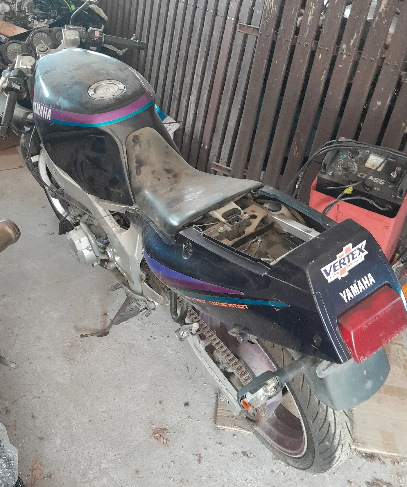 Czarny motocykl marki Yamaha z fioletowymi i niebieskimi akcentami, zaparkowany w garażu na betonowej posadzce. Motocykl jest zakurzony, brakuje mu siedzenia. Na tylnym błotniku widnieją napisy 'VERTEX' oraz 'YAMAHA'. W tle znajduje się drewniany płot z pionowych desek oraz czerwony sprzęt rolniczy z napisem 'CLAAS'.