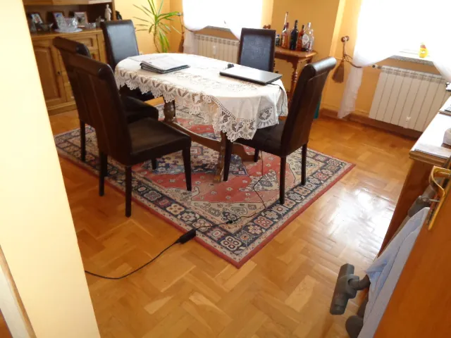 Wnętrze pokoju z jadalnią. W centrum znajduje się owalny stół przykryty białym, koronkowym obrusem, przy którym stoją cztery ciemnobrązowe, skórzane krzesła. Na stole leżą dokumenty i czarny folder. Stół ustawiony jest na ozdobnym dywanie w czerwonych i niebieskich kolorach. Podłoga wykonana jest z drewnianego parkietu ułożonego w jodełkę. W tle widoczny jest drewniany kredens, mały stolik z butelkami alkoholu oraz biały grzejnik pod oknem.