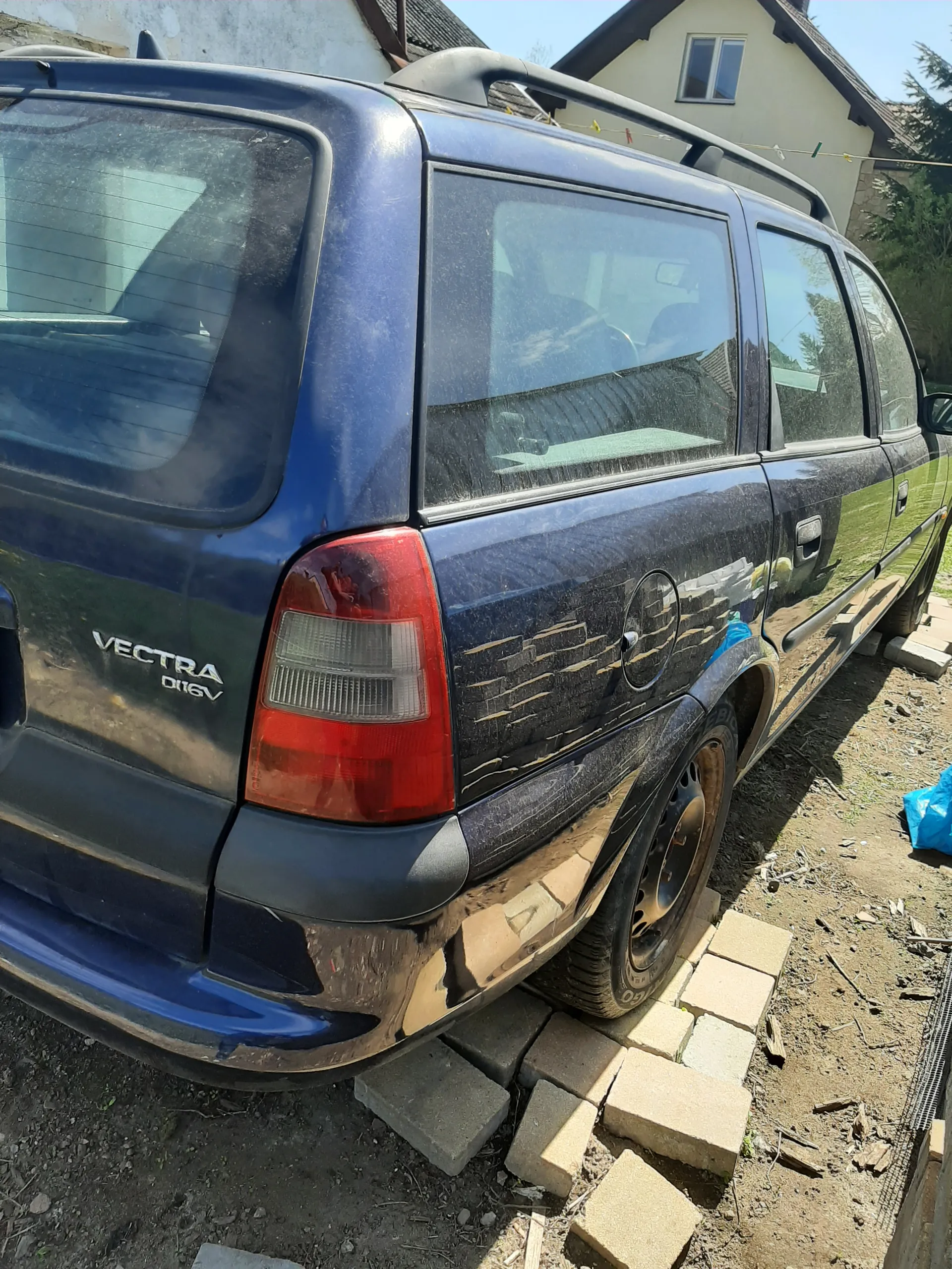 Niebieski samochód osobowy marki VECTRA DI 16V zaparkowany na brudnej powierzchni ziemnej. Widoczne są zarysowane ślady brudu na karoserii, czerwone światło tylnego reflektora oraz odkręcony kołpak koła. W tle znajduje się dom z oknem, stos cegieł i niebieska folia. Na tylnej szybie widoczne są odbicia drzew i budynku.