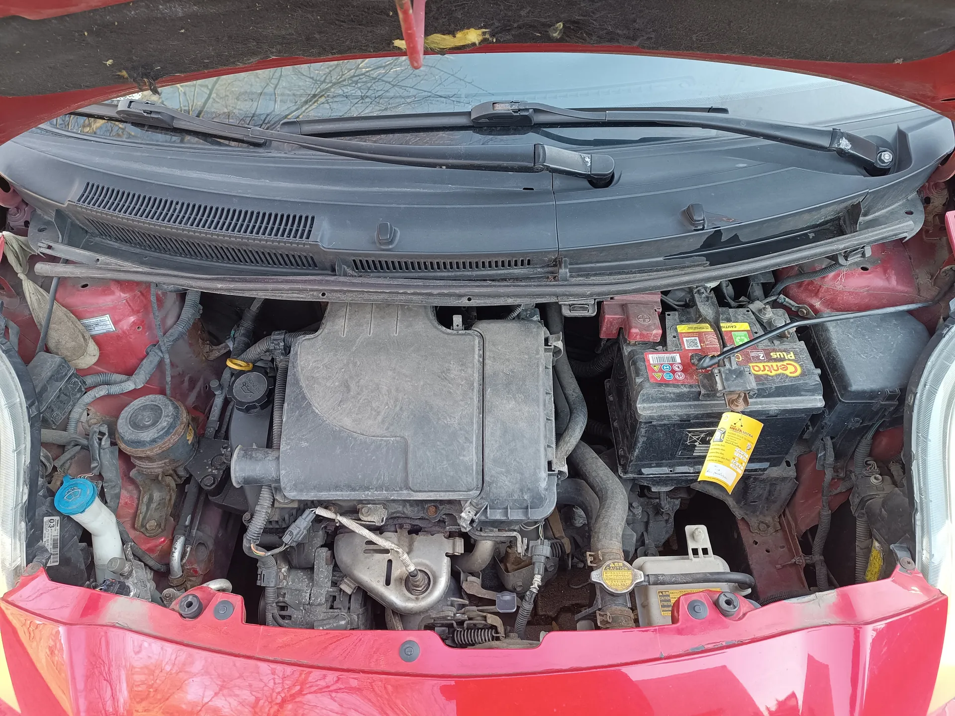 Widok na otwarty maskę czerwonego samochodu z silnikiem. W centralnej części znajduje się zasłona silnika pokryta kurzem. Po prawej stronie widoczna bateria z napisami "Centra" i "Plus 2" oraz żółtą etykietą. Wokół rozłożone są różne elementy, w tym przewody, węże i zbiornik z niebieską nakrętką. Na górze obrazu widoczne są wycieraczki przednie i przednia szyba. Całość pokryta jest kurzem, a czerwona karoseria jest widoczna w dolnej części.