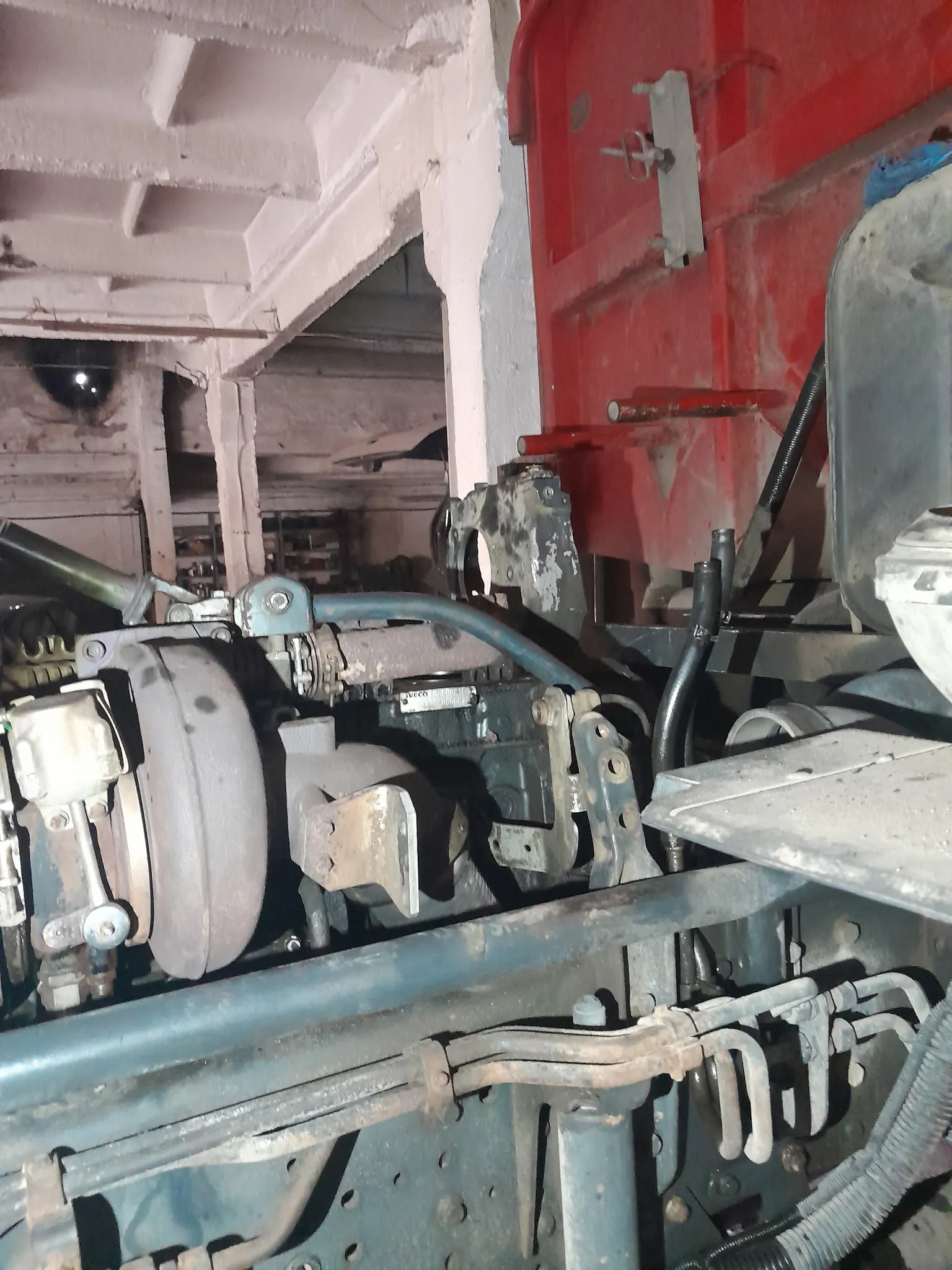 Zbliżenie na złożone elementy mechaniczne, w tym metalowe rury, przewody i części silnika pokryte kurzem oraz oznakami zużycia. W tle widoczna czerwona metalowa konstrukcja oraz białe malowane belki. Na jednej z części silnika znajduje się tabliczka z napisem 'IVECO' oraz cyframi '8034.25'.