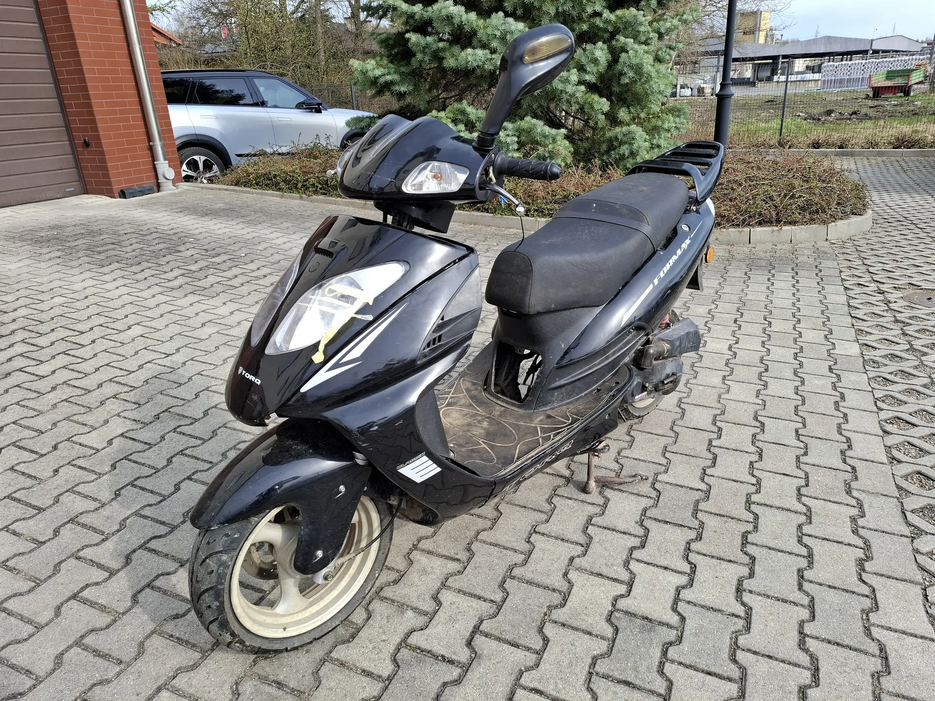 Czarny skuter marki TORO z napisem FORZA na boku, zaparkowany na chodniku z kostki betonowej. W tle srebrny samochód osobowy, ceglany budynek, krzewy i drzewa iglaste. Na skuterze widoczne są reflektory, lusterko wsteczne oraz brudna podłoga. W oddali ogrodzenie i przemysłowe budynki.