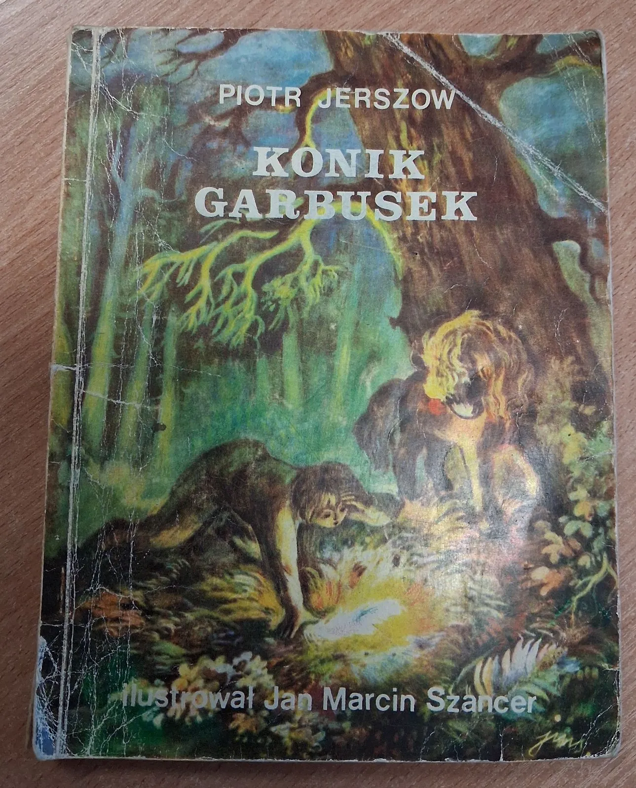 Okładka książki pt. 'KONIK GARBUSEK' autorstwa Piotra Jerszowa, ilustrowana przez Jana Marcina Szancera. Na ilustracji widać dwa konie w gęstym, zielonym lesie pod dużym drzewem. Jeden z koni stoi, a drugi jest pochylony nad jasnym źródłem światła na ziemi. Tekst na górze brzmi: 'PIOTR JERSZOW KONIK GARBUSEK', a na dole: 'ilustrował Jan Marcin Szancer'.