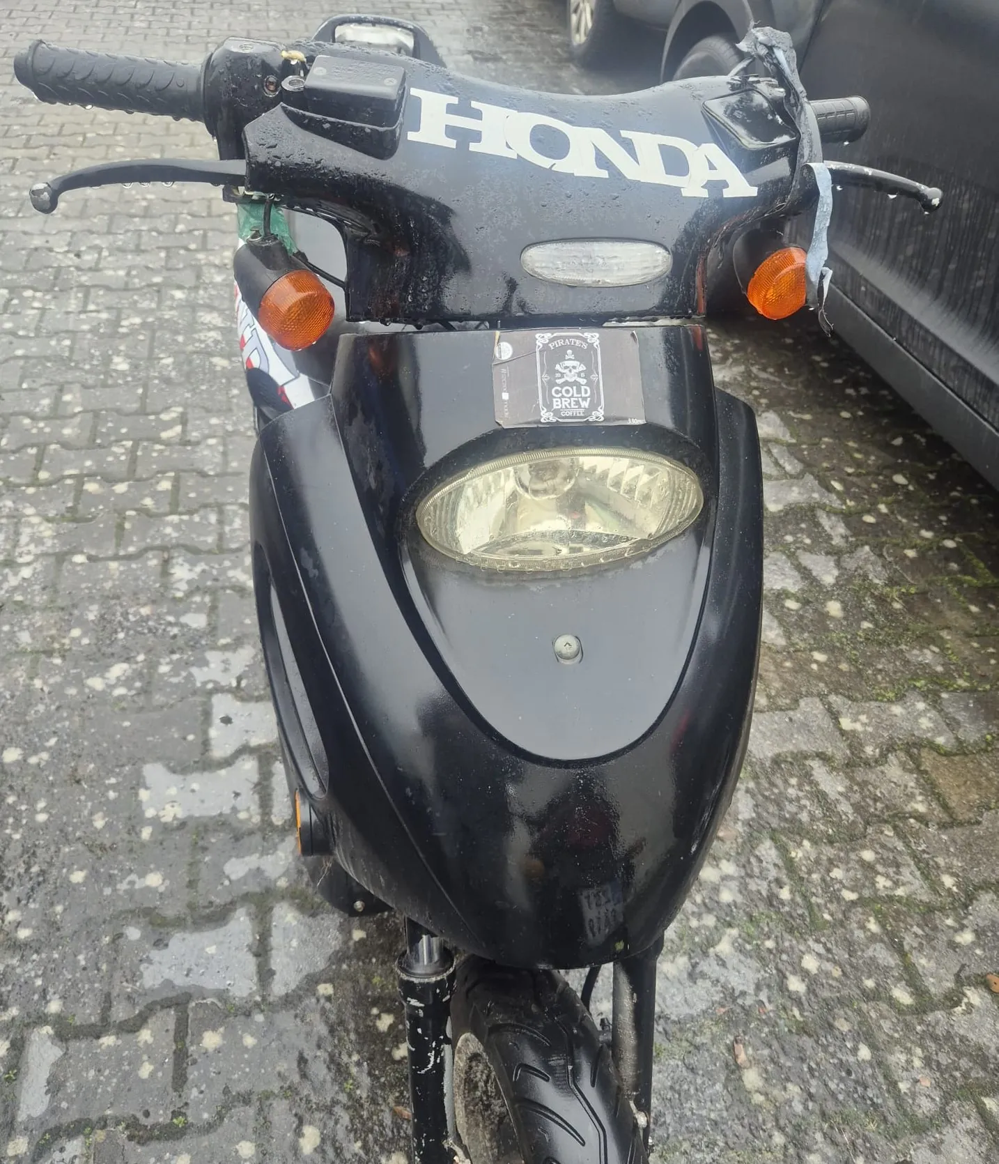 Czarny skuter marki Honda widziany od przodu. Na masce widoczny biały napis HONDA, okrągły reflektor, pomarańczowe kierunkowskazy po obu stronach oraz naklejka z napisem PIRATES COLD BREW COFFEE. Skuter stoi na mokrej, brukowanej nawierzchni. Po prawej stronie częściowo widoczny czarny samochód. Uchwyt kierownicy z czarną obudową i elementami sterującymi.