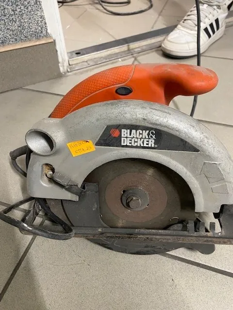 Cyrkularna piła marki BLACK & DECKER o pomarańczowej rękojeści i srebrnym korpusie, z widocznym ostrzem i żółtym etykietą. Piła leży na podłodze z białymi płytkami. W tle widoczna część białego buta z czarnymi paskami oraz przewody elektryczne.