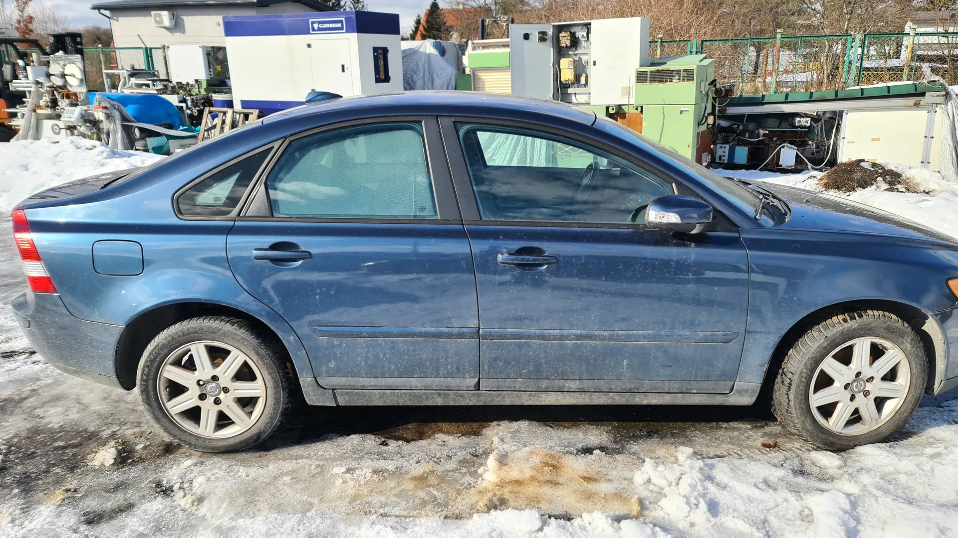 Niebieski sedan zaparkowany na zasypanej śniegiem i lodem powierzchni. Samochód jest zabrudzony, widoczne ślady błota na dolnej części nadwozia. W tle przemysłowe urządzenia, w tym biało-niebieski kontener z napisem "CORMAK", zielone i szare szafy elektryczne oraz inne maszyny. Na ziemi śnieg i odsłonięte fragmenty ziemi. Drzewa bez liści i ogrodzenie w tle.