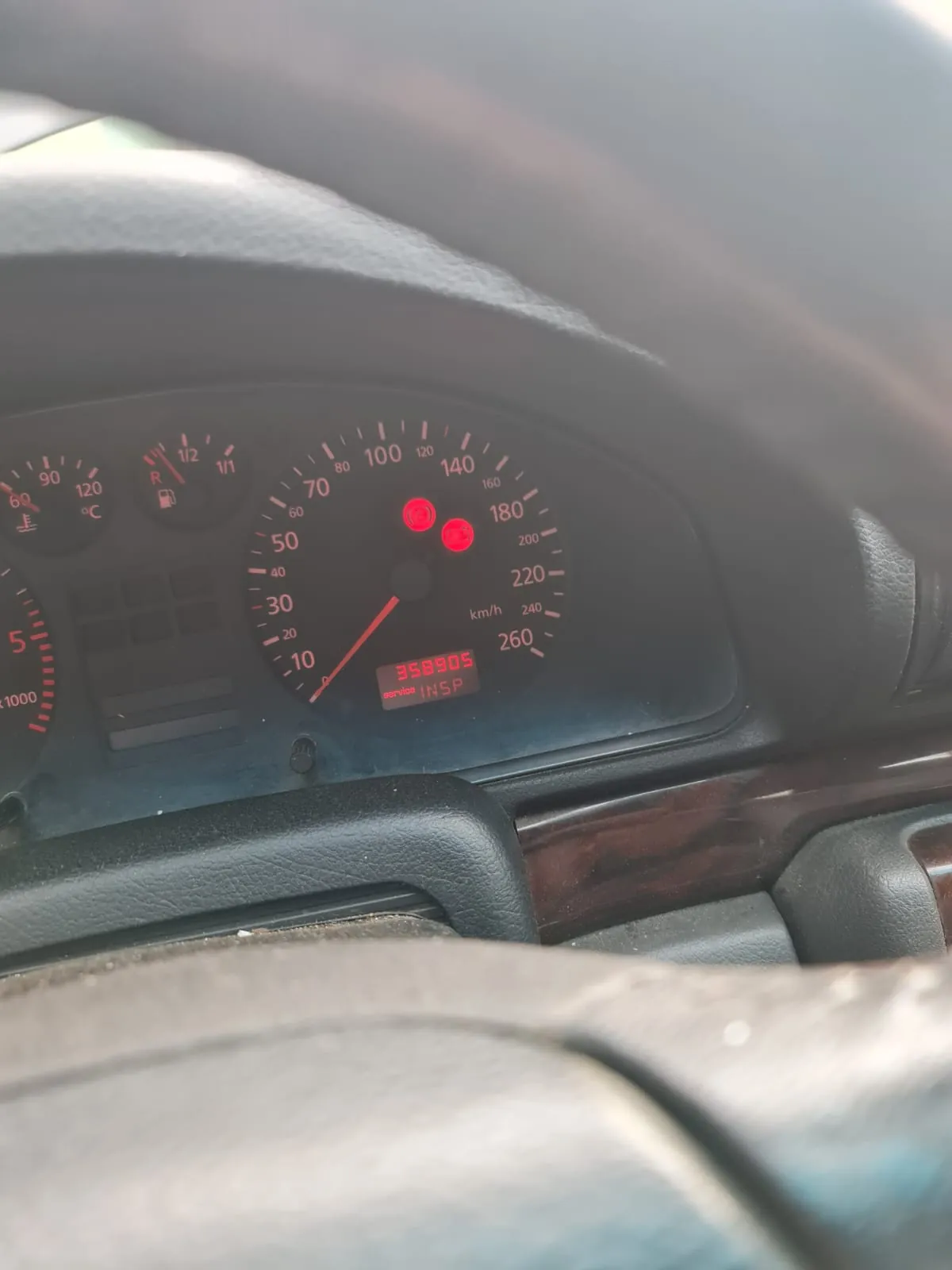 Zbliżenie na deski rozdzielcze samochodu. Widoczny licznik prędkości z zakresem do 260 km/h, obrotomierz, wskaźnik paliwa (blisko zera), termometr (zakres do 120°C) oraz cyfrowy przebieg 358905. Na wyświetlaczu czerwony napis INSP i dwa czerwone lampki ostrzegawcze. Fragment kierownicy w dolnej i górnej części kadru. Deska rozdzielcza z ciemnym tłem i czerwonymi oznaczeniami.