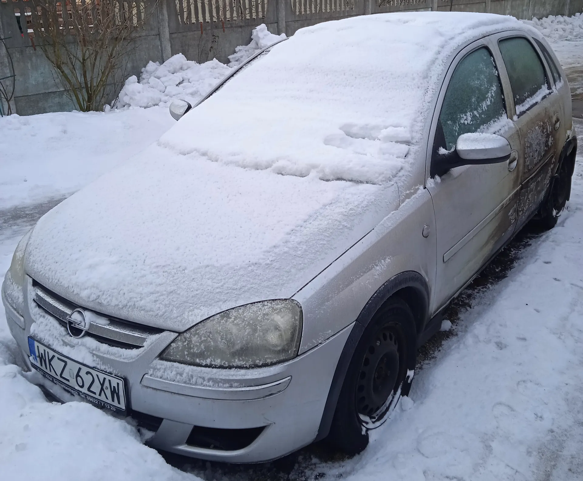 Srebrny samochód marki Opel pokryty grubą warstwą śniegu, zaparkowany na śnieżnej powierzchni. Widoczna tablica rejestracyjna WKZ 62XW oraz logo Opel na masce. W tle betonowy mur z drewnianym ogrodzeniem, nagie krzewy i wysokie kopce śniegu. Samochód ma zabrudzone boki, a przednie światła są częściowo zakryte śniegiem.