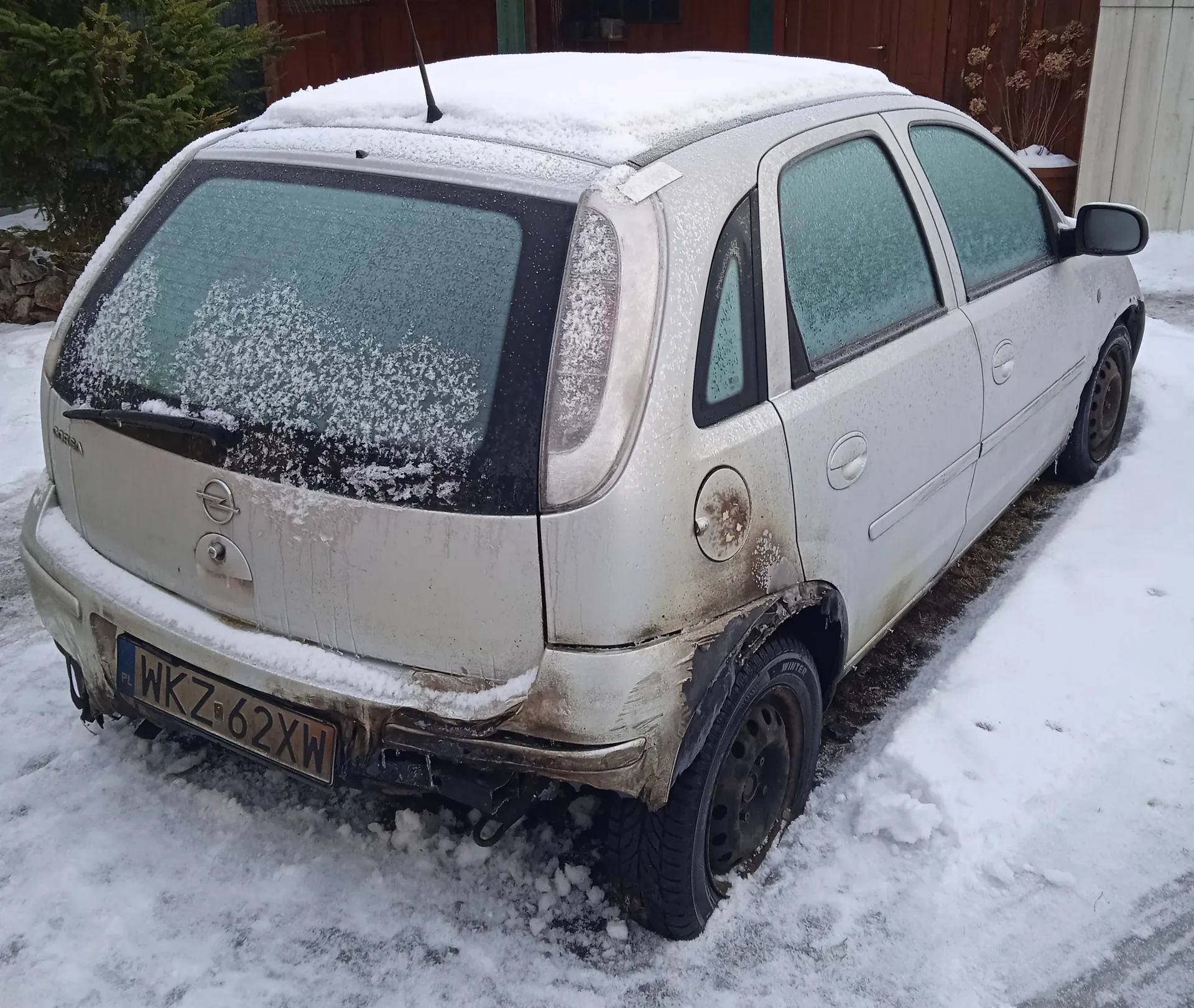 Srebrny samochód marki Opel Corsa pokryty warstwą śniegu, zaparkowany na śnieżnej powierzchni. Widoczna tablica rejestracyjna z napisem WKZ 62XW oraz oznaczeniem PL po lewej stronie. Samochód ma zasłonięte śniegiem szyby, a na karoserii znajdują się ślady brudu. W tle drewniana budowla i zasłonięte śniegiem krzewy. Śnieg pokrywa dach, tylną szybę i fragmenty pasa przedniego.