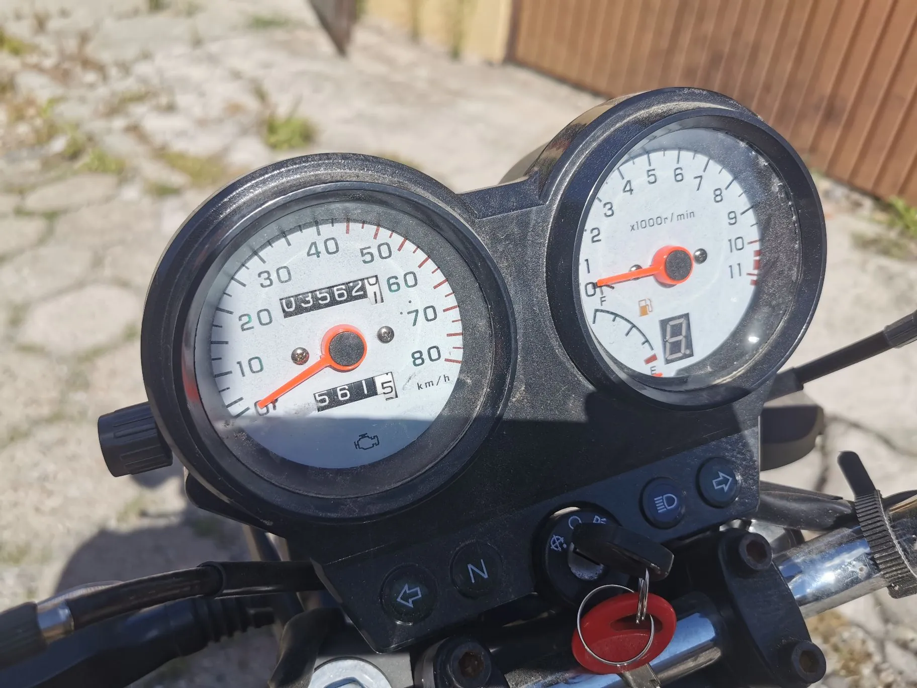 Zbliżenie na deskę rozdzielczą motocykla z dwoma analogowymi wskaźnikami. Lewy wskaźnik to prędkościomierz z podziałką do 80 km/h, z licznikiem przebiegu pokazującym wartość 03562 oraz mniejszym licznikiem poniżej z wartością 5615. Prawy wskaźnik to obrotomierz z podziałką do 11 x1000 r/min. Poniżej wskaźników znajduje się czarny panel z przełącznikami, w tym włącznikiem świateł i stacyjką z włożonym kluczykiem z czerwonym brelokiem.