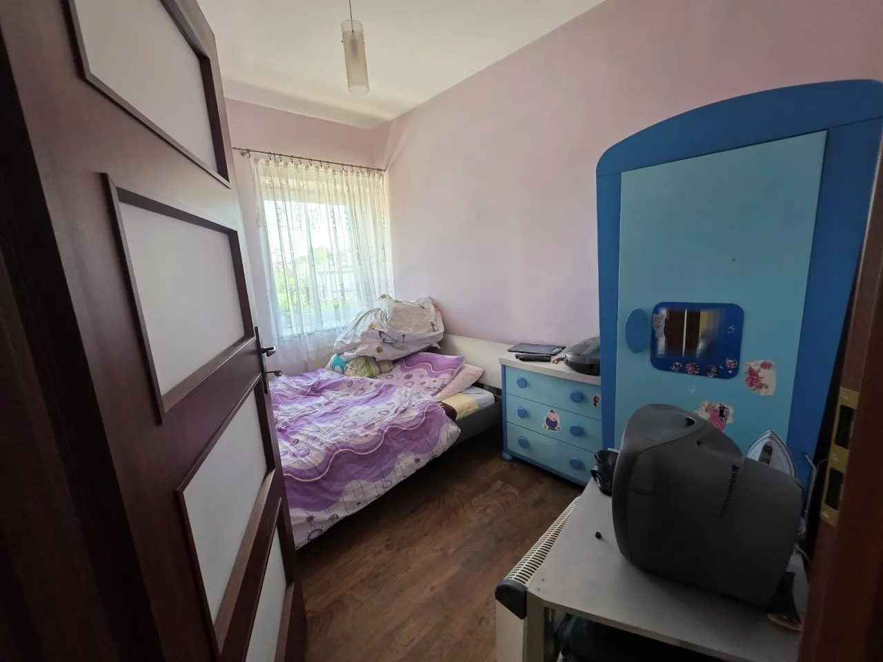 Widok z otwartych drzwi do małego pokoju z jasnofioletowymi ścianami. W pomieszczeniu znajduje się łóżko z fioletową pościelą, niebieska komoda z szufladami oraz duża niebieska szafa z lustrem. Na pierwszym planie po prawej stronie stoi biały stolik z ciemnoszarym monitorem CRT. W tle widoczne jest okno z białymi firankami i wisząca lampa sufitowa.