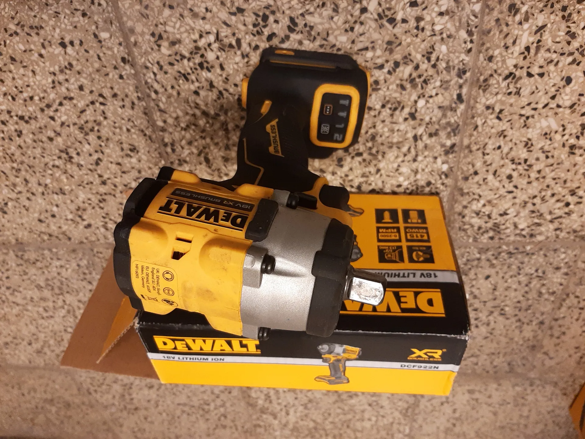 Żółto-czarny akumulatorowy wkrętarko-wkrętarka DeWALT 18V XR Brushless z widocznym wyświetlaczem baterii pokazującym wartość 21. Za narzędziem znajduje się otwarte pudełko z logo DeWALT, napisem 18V LITHIUM oraz specyfikacjami: 415 MWO, 0-2800 RPM. Tło stanowi posadzka z kruszywem w kolorze beżowym z czarnymi i białymi kropkami.