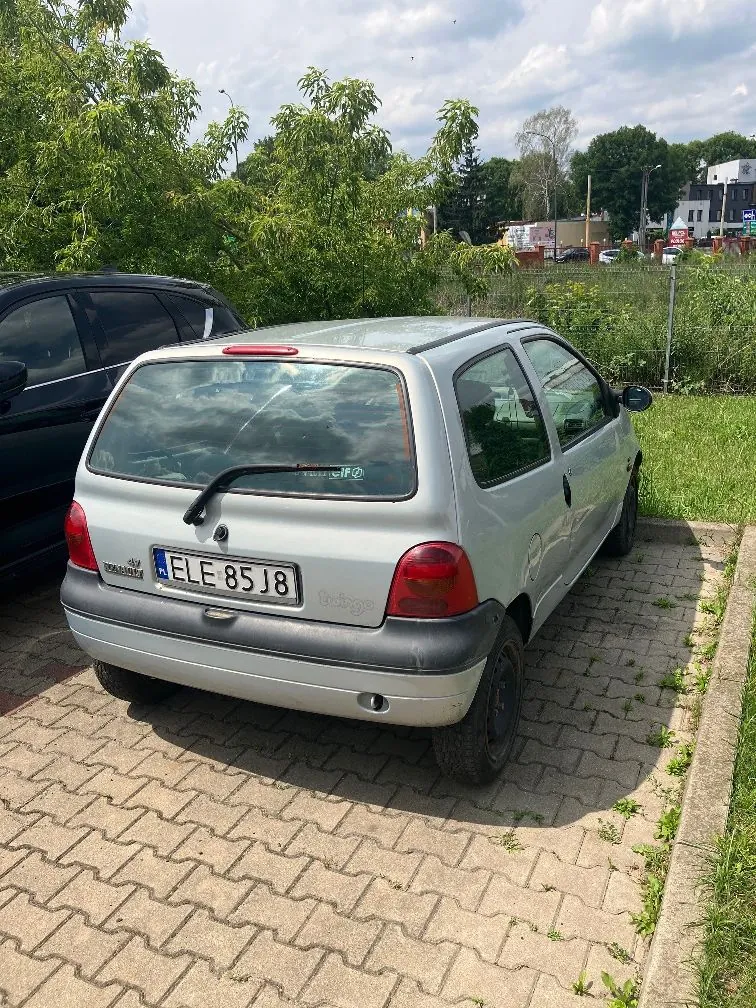 Srebrny samochód marki Renault Twingo zaparkowany na kostce brukowej. Widoczna jest tylna część pojazdu z tablicą rejestracyjną o numerze ELE 85J8. Obok stoi czarny samochód, a w tle znajdują się zielone drzewa, krzewy oraz fragmenty zabudowań pod zachmurzonym niebem.