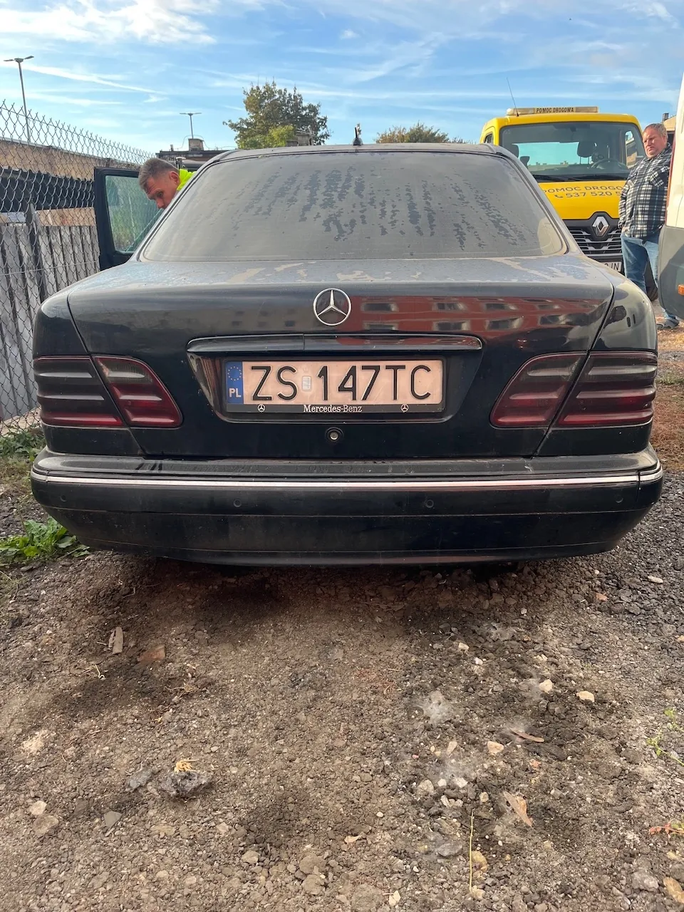 Tylny widok czarnego samochodu marki Mercedes-Benz z tablicą rejestracyjną ZS 147TC. Samochód stoi na nieutwardzonym, kamienistym podłożu. W tle widoczny jest żółty samochód pomocy drogowej oraz mężczyzna stojący obok niego.