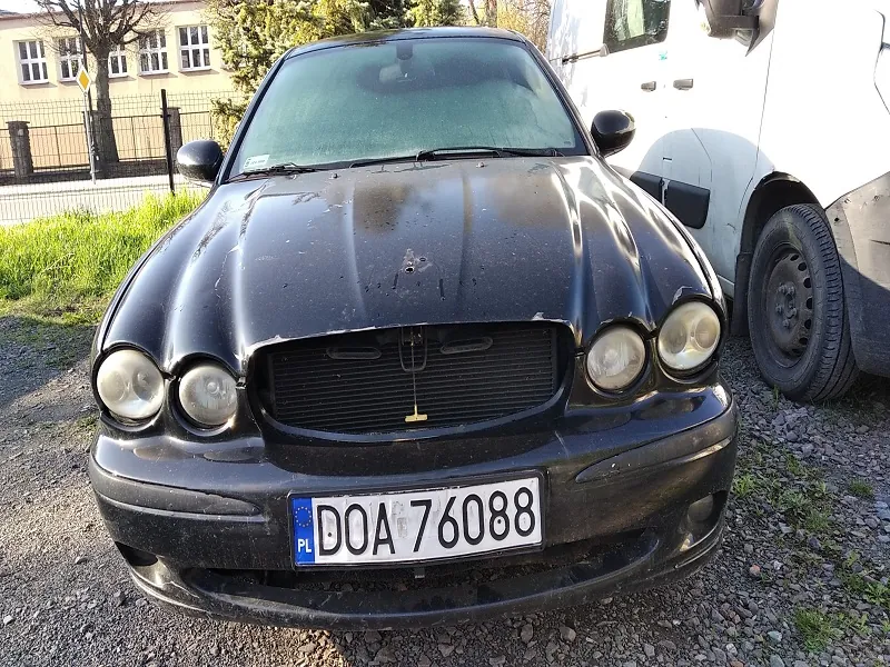 Czarny samochód osobowy marki Jaguar z przodu, z widoczną tablicą rejestracyjną PL DOA 76088. Samochód stoi na żwirowym podłożu, obok częściowo widoczny biały pojazd ciężarowy. W tle budynek z oknami, ogrodzenie, trawa i drzewa. Samochód jest zabrudzony, a przednia maska ma ślady uszkodzeń. Słońce oświetla scenę, tworząc cienie na ziemi.