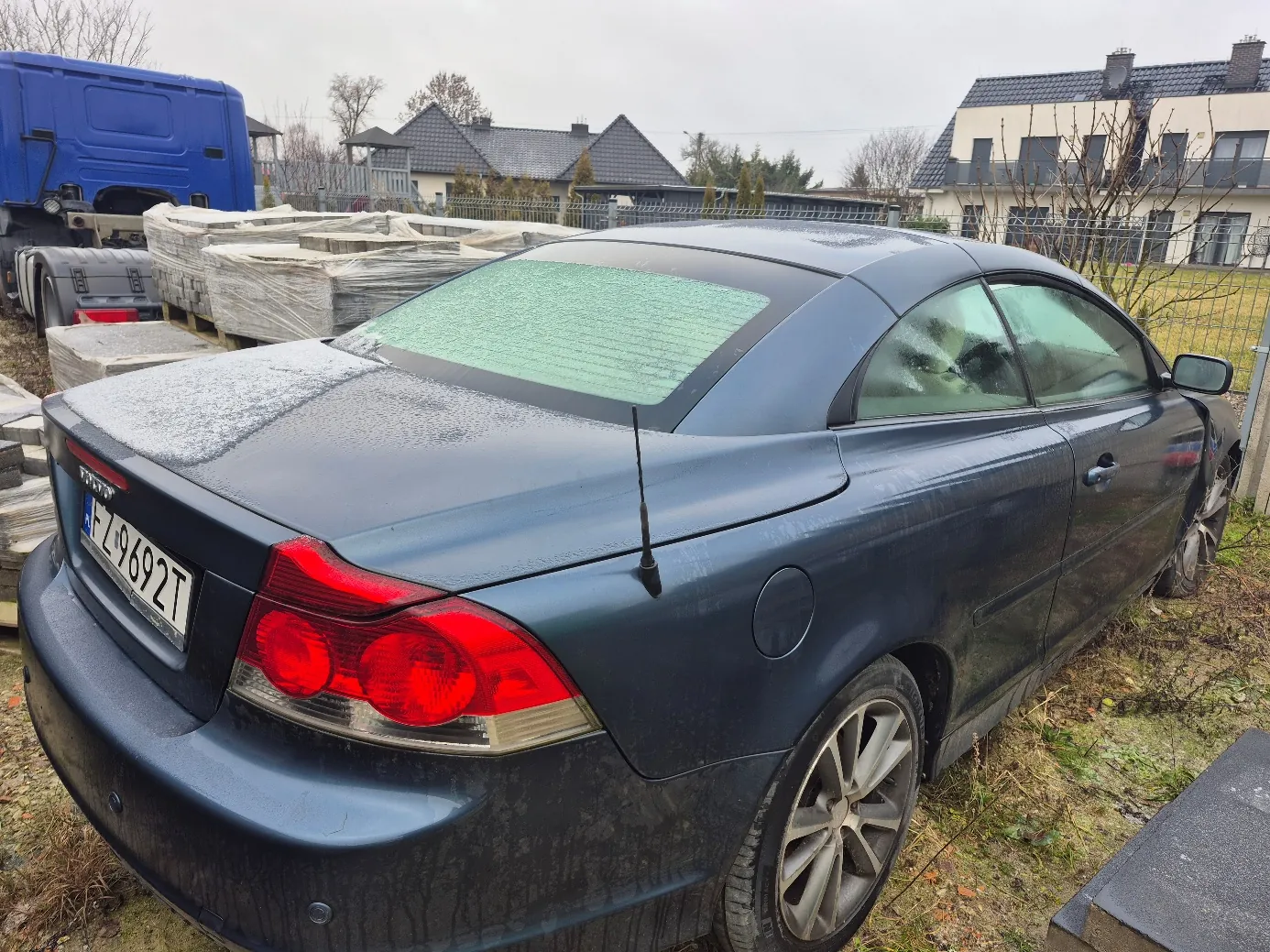 Ciemnoniebieski samochód Audi coupe z tablicą rejestracyjną FZ 9692T, zaparkowany na trawie. Na dachu i tylnym szybie widoczny jest śnieg lub szron. W tle niebieska ciężarówka, stosy materiałów owiniętych folią, domy z ciemnymi dachami oraz ogrodzenie. Niebo jest zachmurzone, a trawa na terenie jest częściowo zasłana suchymi liśćmi.