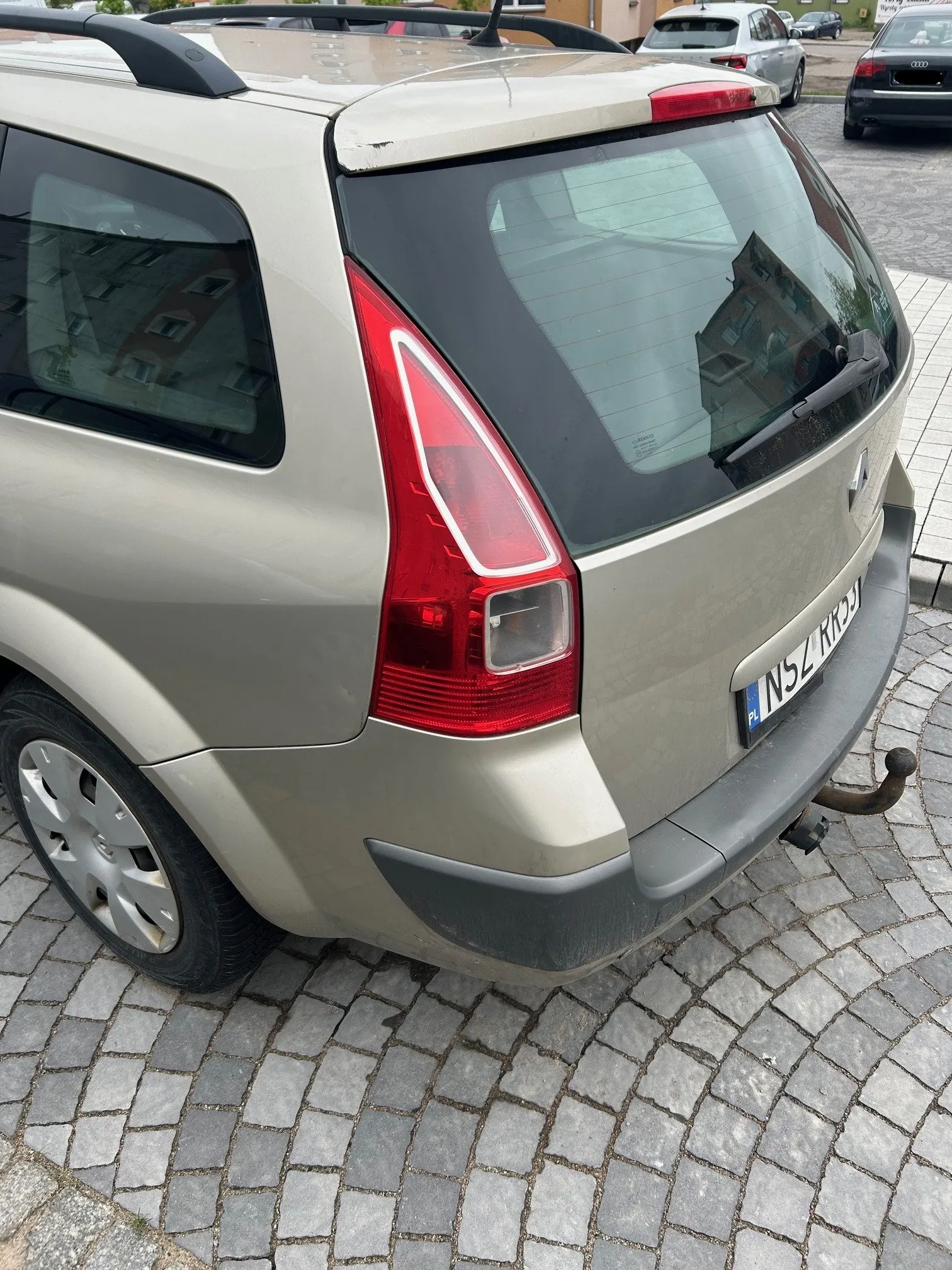 Tył beżowego samochodu typu SUV zaparkowanego na brukowanej nawierzchni. Widoczne są czerwone światła tylnego, tablica rejestracyjna z napisem PL NSZ RRSJ, hak holowniczy oraz refleksje budynków w szybie tylnej. W tle stoją inne pojazdy, w tym srebrny i czarny samochód. Samochód ma dachowy bagażnik i srebrne felgi.