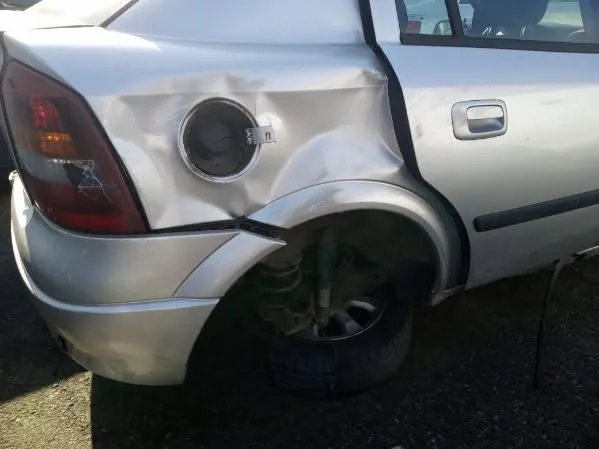 Zdjęcie przedstawia tylną część srebrnego samochodu z widocznymi uszkodzeniami. Lewe tylne nadkole jest zdeformowane, a koło zostało usunięte, odsłaniając zawieszenie. Otwarty klosz zbiornika paliwa znajduje się na boku pojazdu. Samochód stoi na podłożu z drobnej kostki. W tle widoczne są fragmenty innych pojazdów.