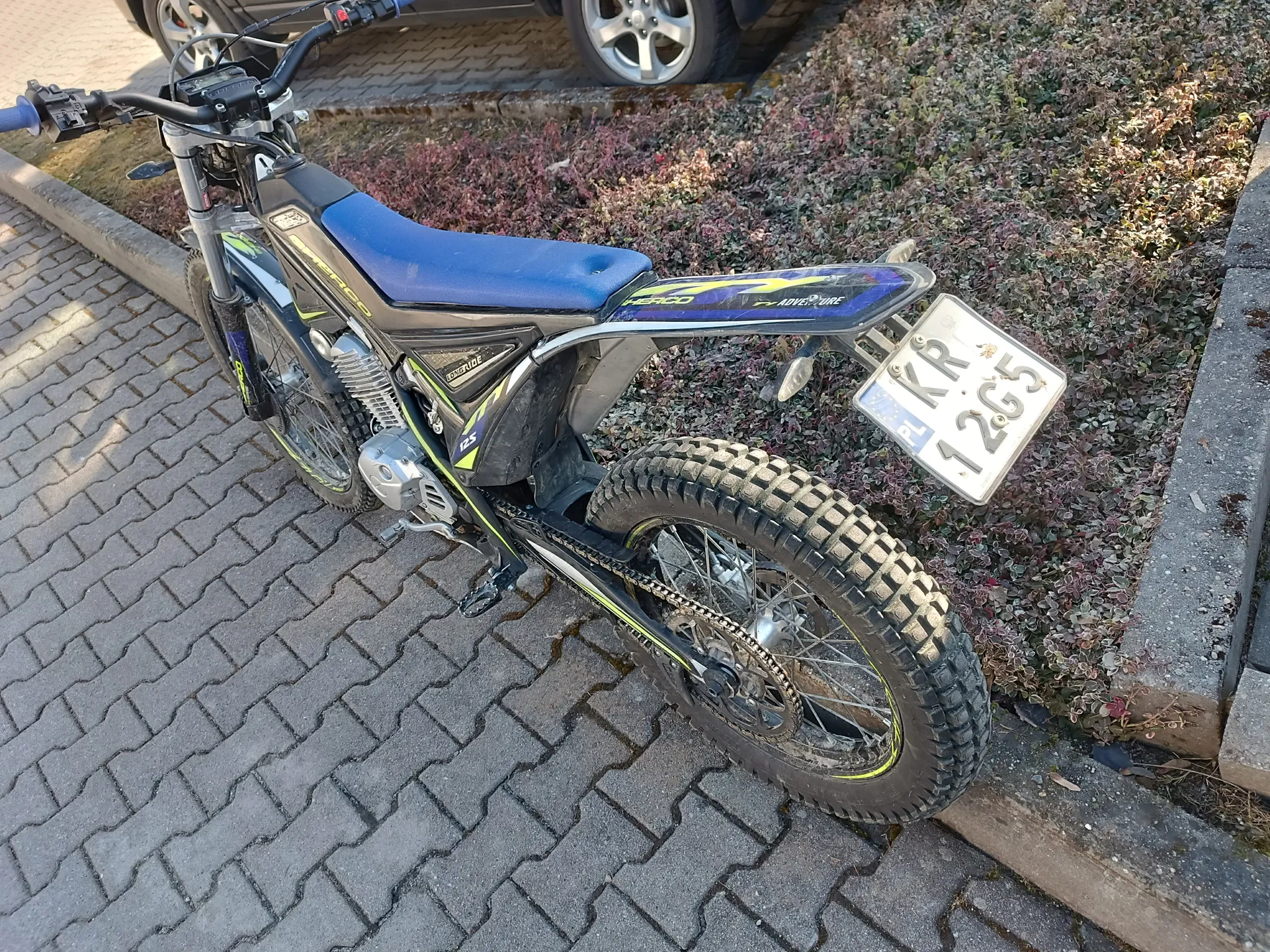 Mały motocykl enduro czarny z żółtymi i niebieskimi akcentami, niebieskim siedzeniem oraz tablicą rejestracyjną PL KR 12G5. Pojazd stoi na brukowanej nawierzchni obok chodnika i krzewów o brązowo-zielonych liściach. W tle widoczne są koła samochodu.