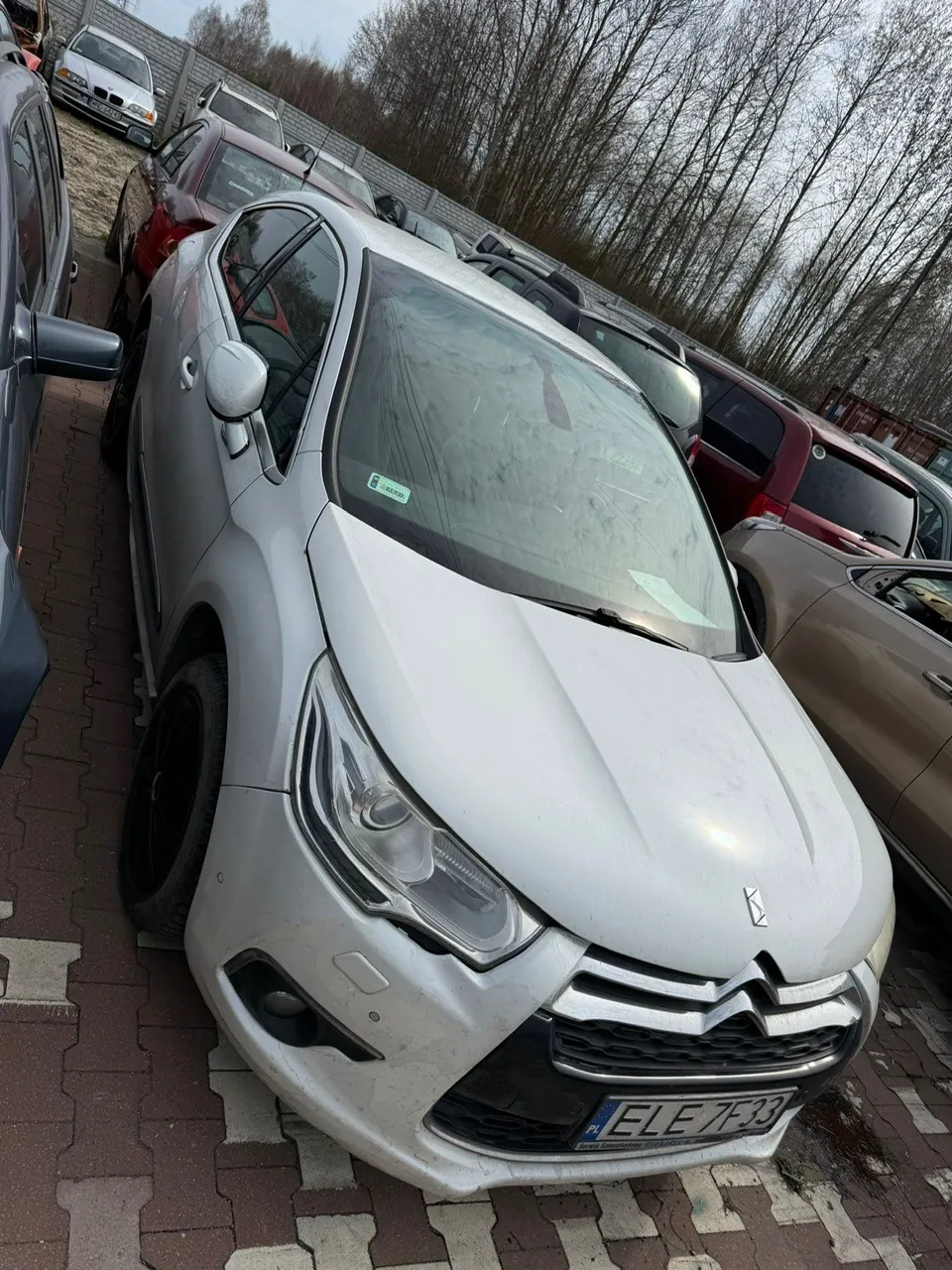 Srebrny samochód Citroën z tablicą rejestracyjną ELE 7F33 zaparkowany na placu z czerwono-białymi płytami. W tle inne pojazdy, drzewa bez liści i ogrodzenie. Na przedniej szybie widoczny zielony znaczek. Samochód znajduje się w centrum kadru, otoczony innymi autami po obu stronach.