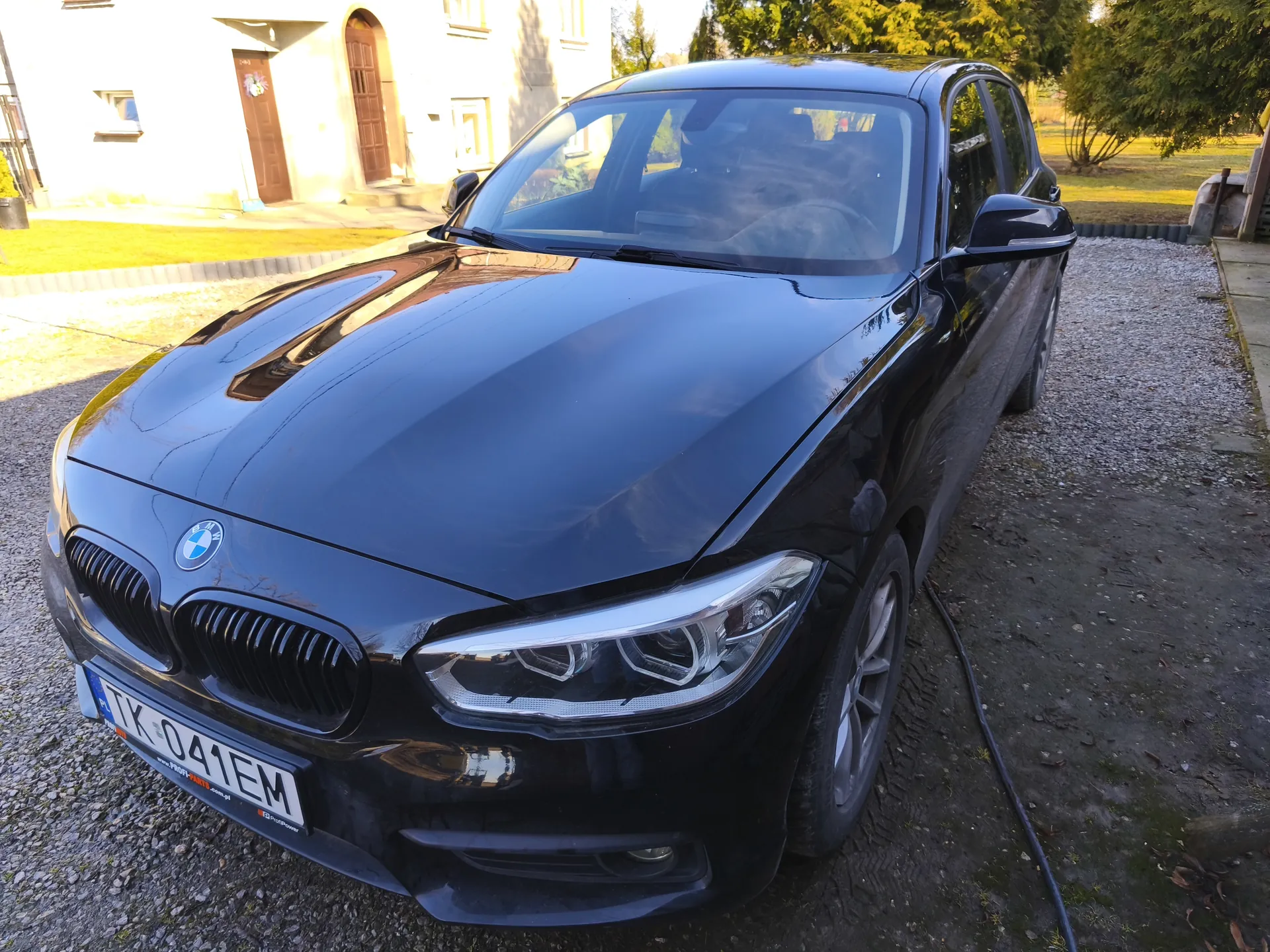 Czarny samochód marki BMW zaparkowany na podjeździe z żwiru. Widoczna tablica rejestracyjna z napisem TK 041EM. W tle budynek z brązowymi drzwiami i oknami, trawnik oraz drzewa. Samochód ma charakterystyczne podwójne klapki chłodnicy i logo BMW na masce. Przy samochodzie leży czarny przewód.