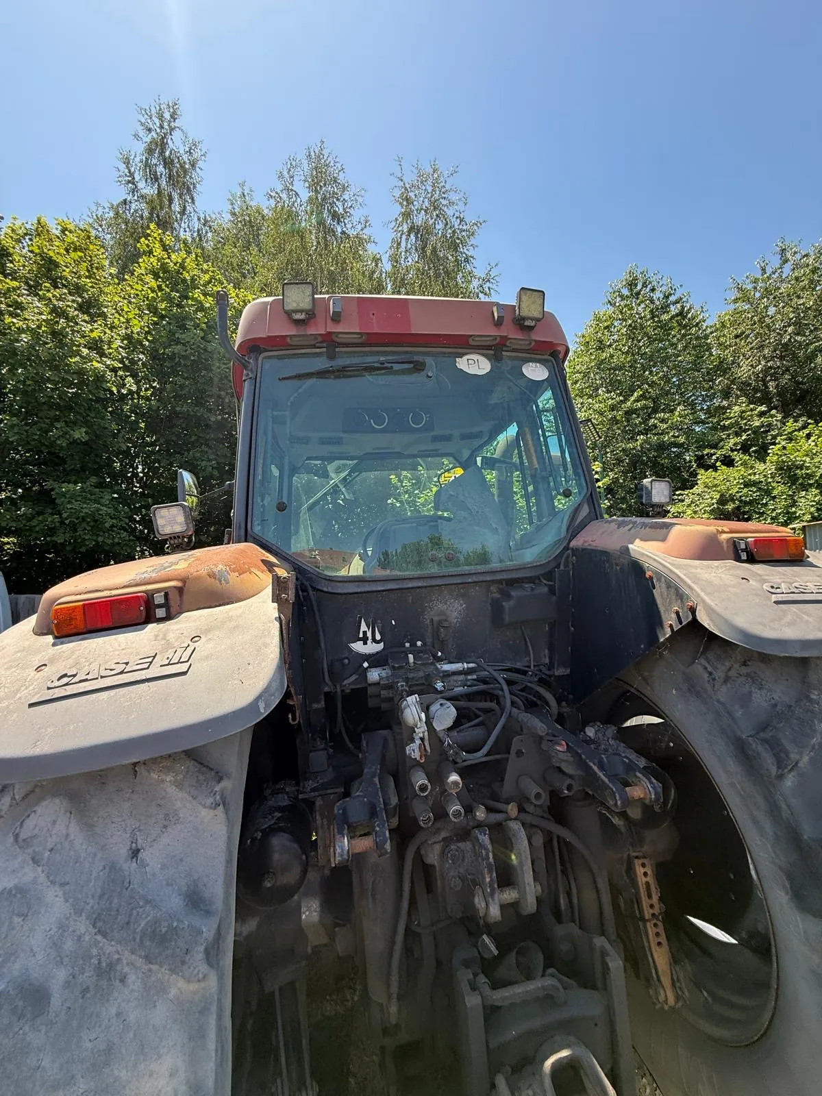 Widok z przodu na czerwony traktor marki Case IH. Widoczna jest kabina z przeszkloną szybą, reflektory na dachu oraz przedni układ hydrauliczny z licznymi przewodami i złączami. Traktor stoi na tle zielonych drzew pod bezchmurnym, niebieskim niebem.