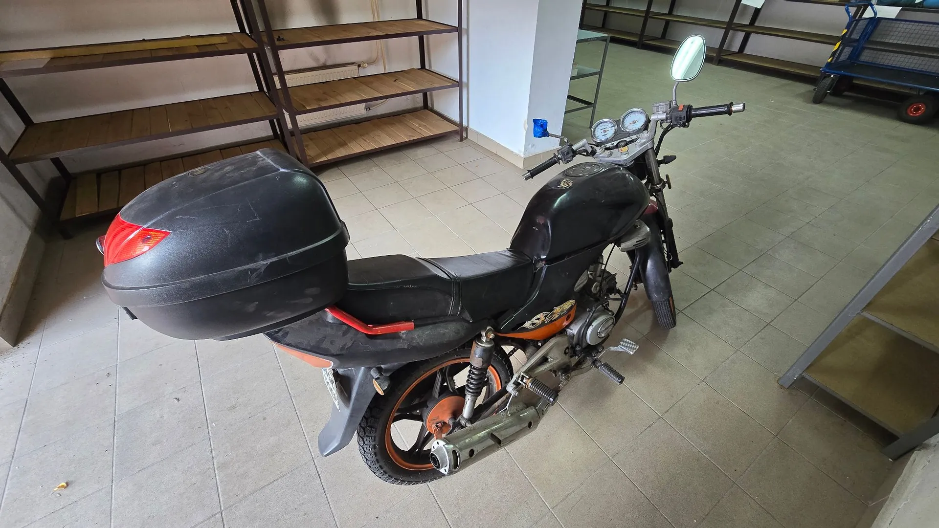 Czarny motocykl zaparkowany w pomieszczeniu z jasną kafelkową podłogą. Motocykl posiada zamontowany duży, czarny kufer z czerwonym światłem tylnym. W tle widoczne są drewniane regały magazynowe oraz fragment niebieskiego wózka transportowego.