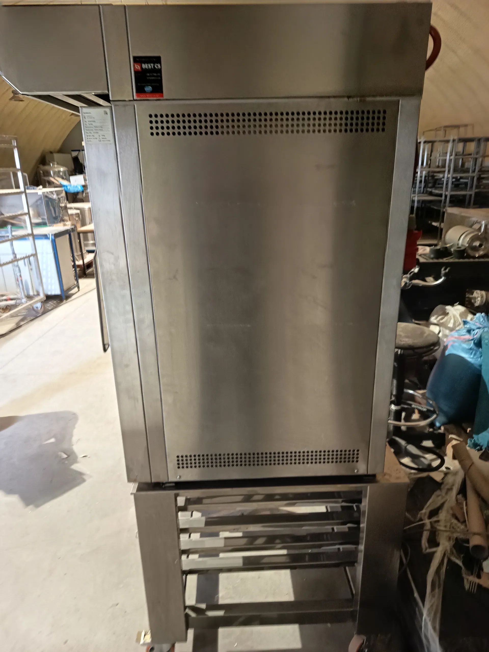 Wysoki, stalowy przemysłowy aparat z perforowanymi otworami na górnej i dolnej części. Na górze urządzenia widoczna czarna tabliczka z napisem 'BEST CS' oraz dodatkowymi informacjami. W tle warsztat z metalowymi półkami, narzędziami i różnymi przedmiotami. Podłoga z betonu, po prawej stronie widoczne są nieuporządkowane materiały i sprzęt.