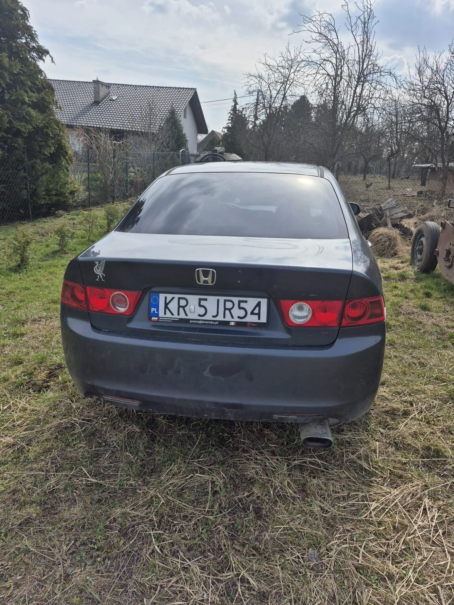 Tył ciemnoszarego samochodu marki Honda z tablicą rejestracyjną KR 5 JR54 (PL po lewej stronie). Na lewej stronie bagażnika naklejka z logo Liverpool FC. Samochód stoi na trawie, w tle dom z dachem krytym dachówką, drzewa, ogrodzenie i stosy siana. Po prawej stronie widoczne są stare opony i metalowe elementy. Niebo częściowo zachmurzone.