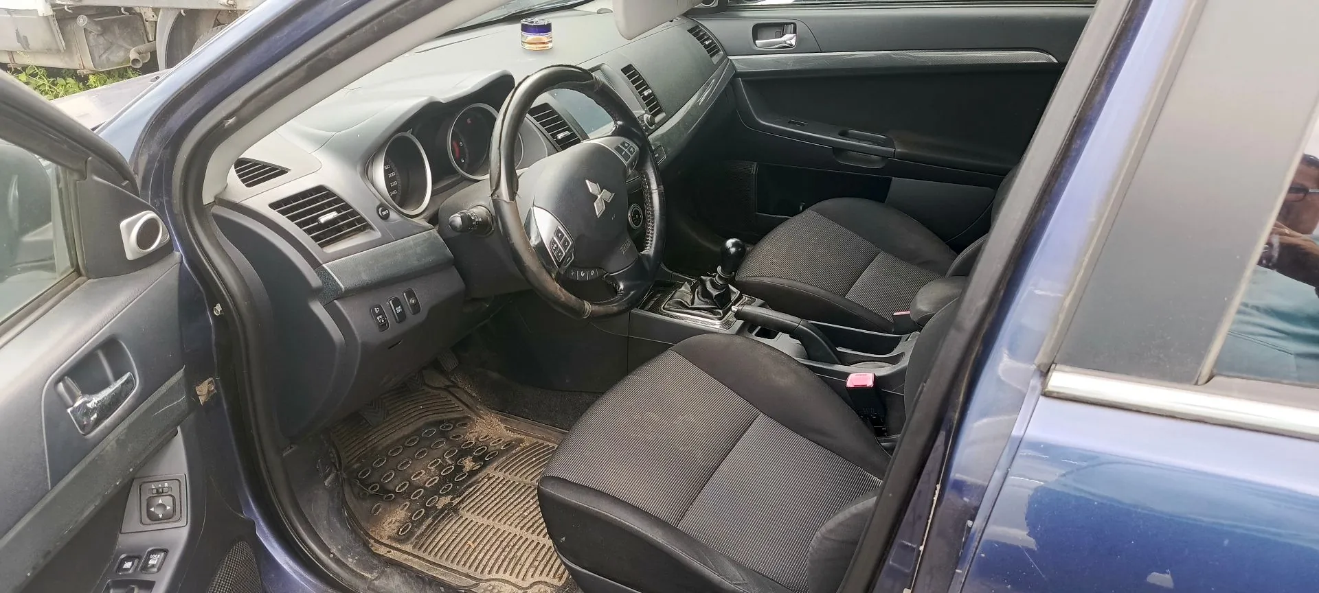 Wnętrze niebieskiego samochodu widziane z otwartych drzwi kierowcy. Widoczna jest czarna tapicerka materiałowa na fotelach, kierownica z logo Mitsubishi, deska rozdzielcza z zegarami, lewarek zmiany biegów oraz gumowa mata na podłodze.