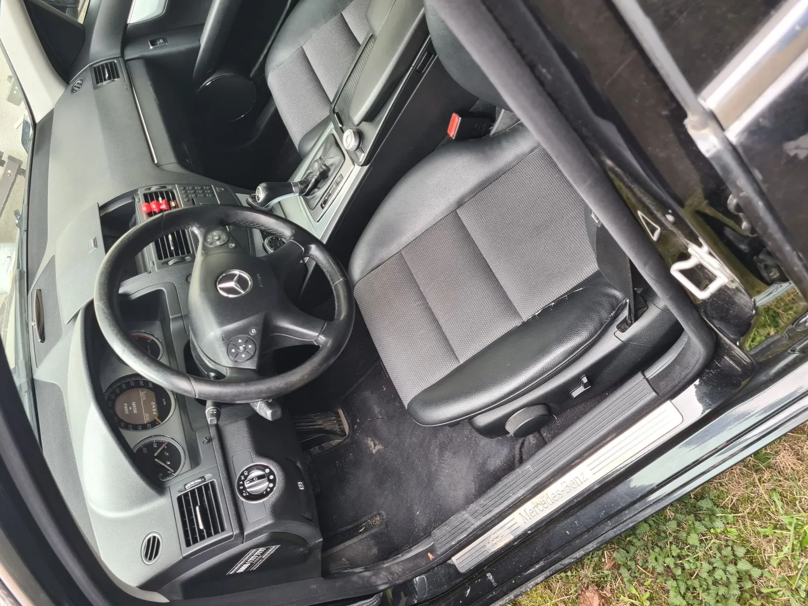 Wnętrze samochodu Mercedes-Benz z widoczną kierownicą z logo, deską rozdzielczą, czarno-szarymi siedzeniami przodem, dźwignią zmiany biegów, otwartymi drzwiami i napisem „Mercedes-Benz” na progu. W tle trawa.