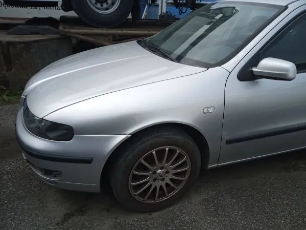 Srebrny samochód osobowy typu sedan, widok z przodu i boku. Widoczna przednia część z reflektorem, felgami aluminiowymi oraz fragmentem przedniego pasa. Samochód zaparkowany na nawierzchni asfaltowej. W tle betonowe bloki i fragment innego pojazdu z kołem. Brak czytelnego tekstu.