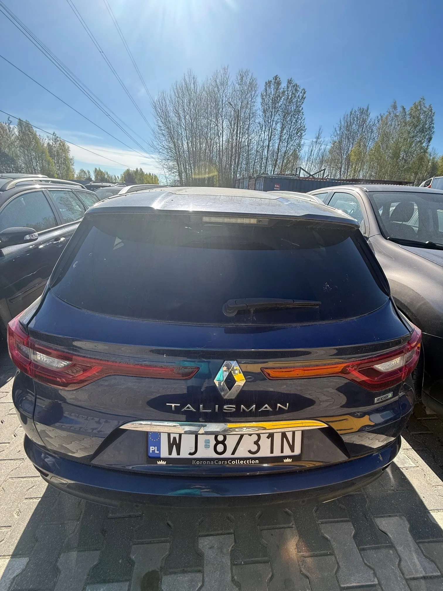 Tył niebieskiego samochodu marki Renault Talisman z widocznym logo, napisem TALISMAN oraz tablicą rejestracyjną PL WJ 8731N. Poniżej tablicy napis Korona Cars Collection. Samochód zaparkowany na placu parkingowym wśród innych pojazdów. W tle drzewa, niebo z słońcem i linie energetyczne.