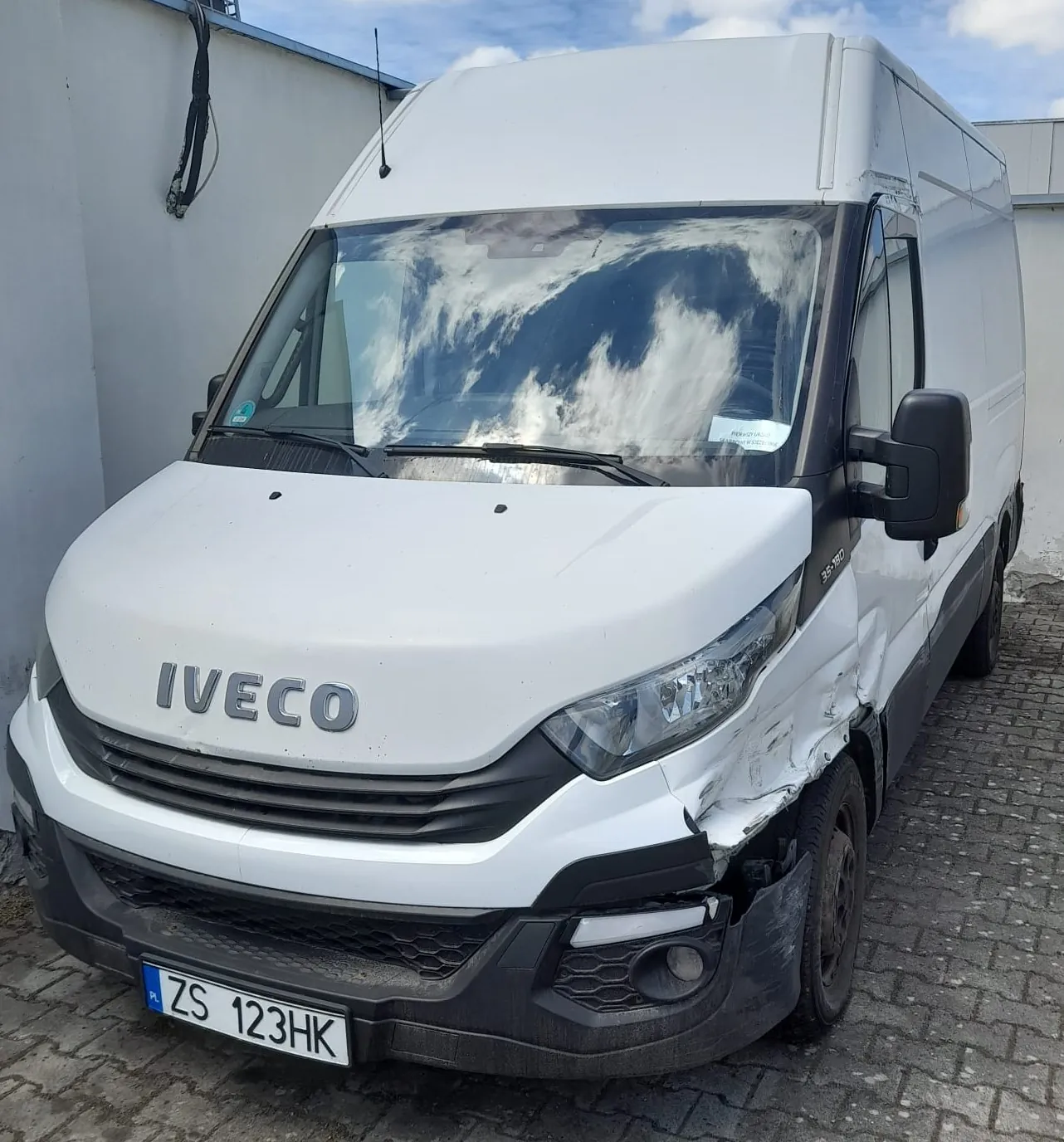 Biały furgon marki Iveco zaparkowany na kostce brukowej. Pojazd ma widoczne uszkodzenia karoserii po prawej stronie przodu, w tym wgniecenie błotnika i poluzowany czarny zderzak. Na masce widnieje napis 'IVECO', a tablica rejestracyjna ma numer 'ZS 123HK'.