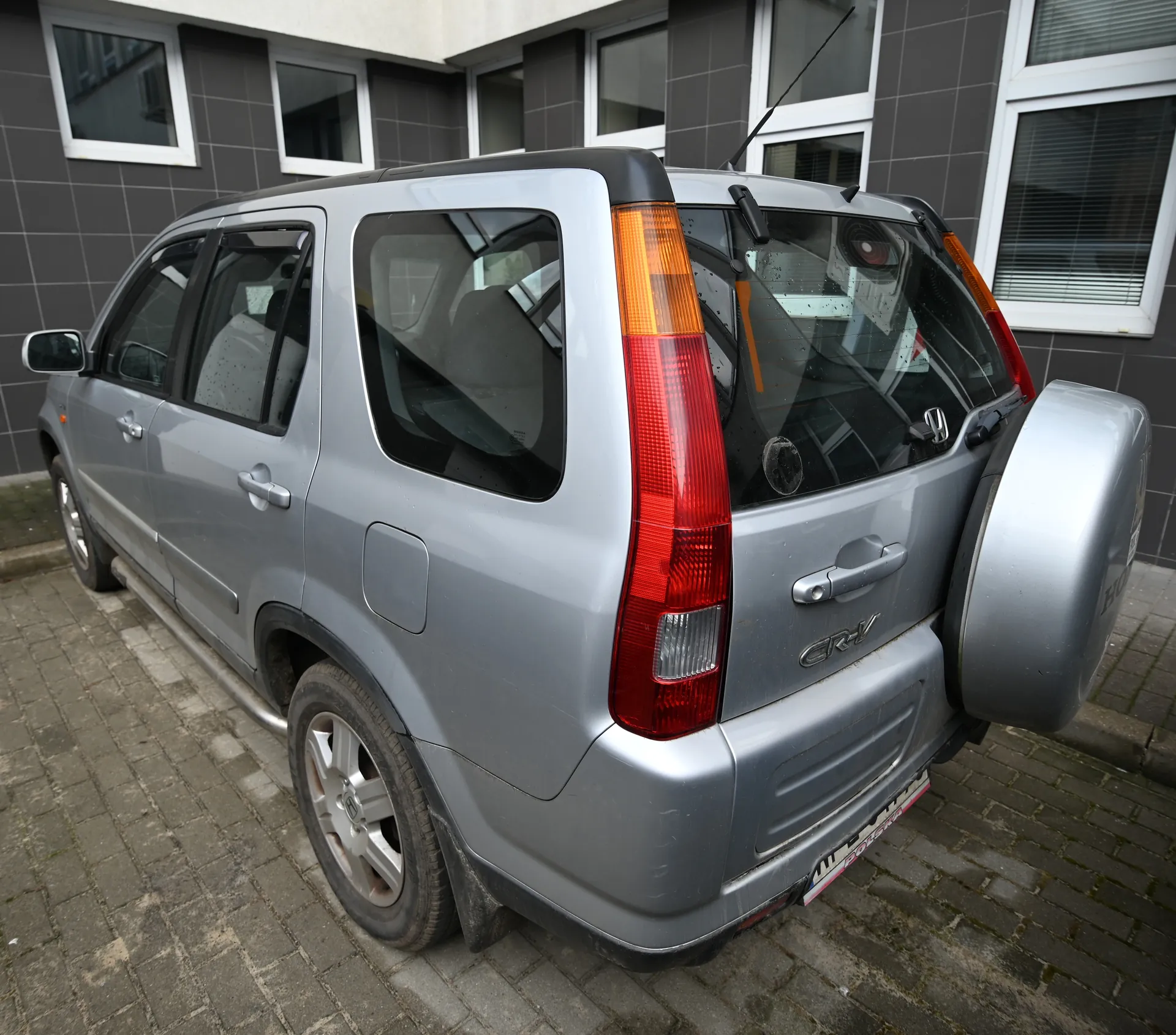 Srebrny samochód typu SUV marki Honda CR-V zaparkowany na brukowanej powierzchni przed budynkiem z szarymi płytkami i białymi oknami. Widoczne są czerwone światła tylne, koło zapasowe na tylnej klapie oraz napis CR-V na bagażniku.