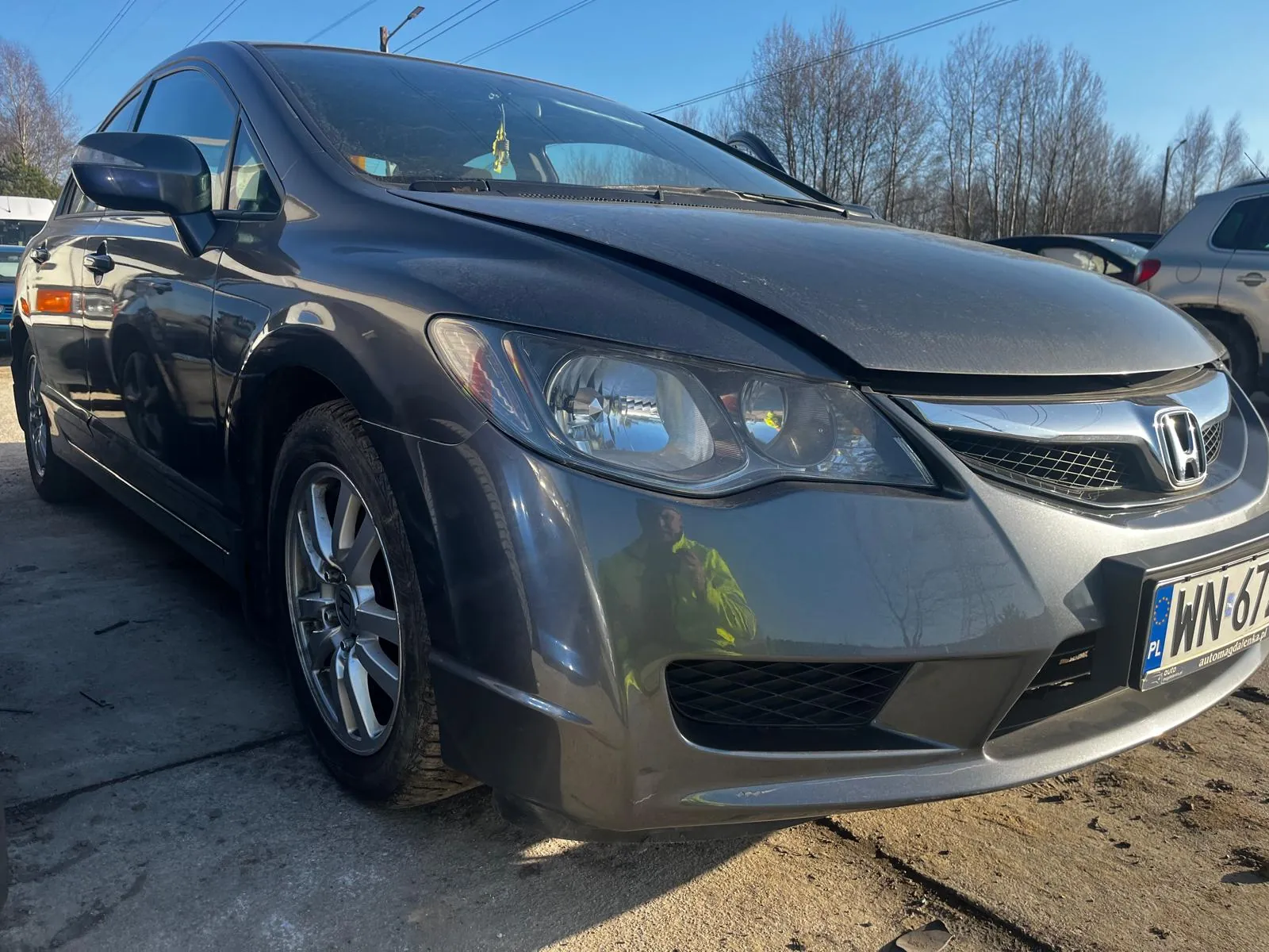 Ciemnoszary samochód marki Honda z widocznym logo na masce, z tablicą rejestracyjną PL WN 67 i częściowo widocznym napisem automagdalenka.pl. Na masce odbicie osoby w żółtej kurtce. Tło: inne zaparkowane pojazdy, drzewa bez liści, linie energetyczne i niebo. Samochód stoi na betonowej i ziemnej powierzchni.