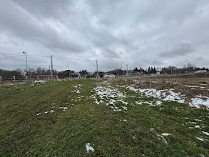 Rozległe pole z zieloną trawą i plamami śniegu pod szarym, zachmurzonym niebem. W tle widoczne są słupy energetyczne, ogrodzenie oraz rozproszone budynki. Horyzont jest poziomy, a całość ujęta w naturalnej perspektywie.