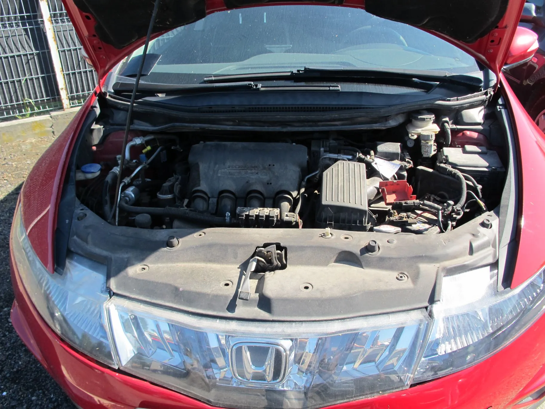 Widok z góry na otwartą maskę czerwonego samochodu marki Honda, ukazującą silnik i elementy komory silnika. W dolnej części zdjęcia widoczny jest przedni pas z reflektorami oraz srebrna atrapa chłodnicy z logo Hondy.