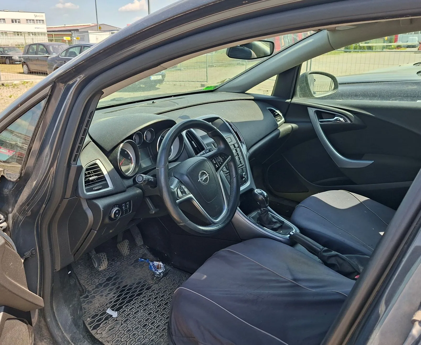 Wnętrze samochodu marki Opel, widok z otwartych drzwi kierowcy. Widoczna jest czarna deska rozdzielcza, kierownica z logo producenta, zegary, konsola środkowa z lewarkiem zmiany biegów oraz dwa przednie fotele pokryte ciemnymi pokrowcami. Na podłodze znajduje się gumowa mata z widocznymi drobnymi zanieczyszczeniami. Przez szyby widoczny jest parking z innymi samochodami i budynkami w tle.