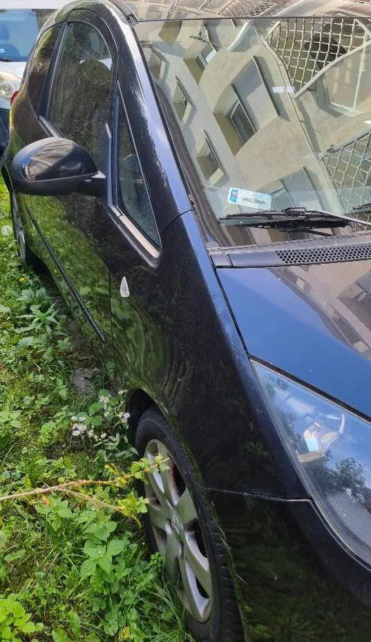 Zbliżenie na prawy przedni bok czarnego samochodu osobowego zaparkowanego przy krawężniku z zieloną roślinnością. Widoczna jest przednia szyba z odbiciem budynku, lusterko boczne oraz przednie koło z felgą aluminiową.