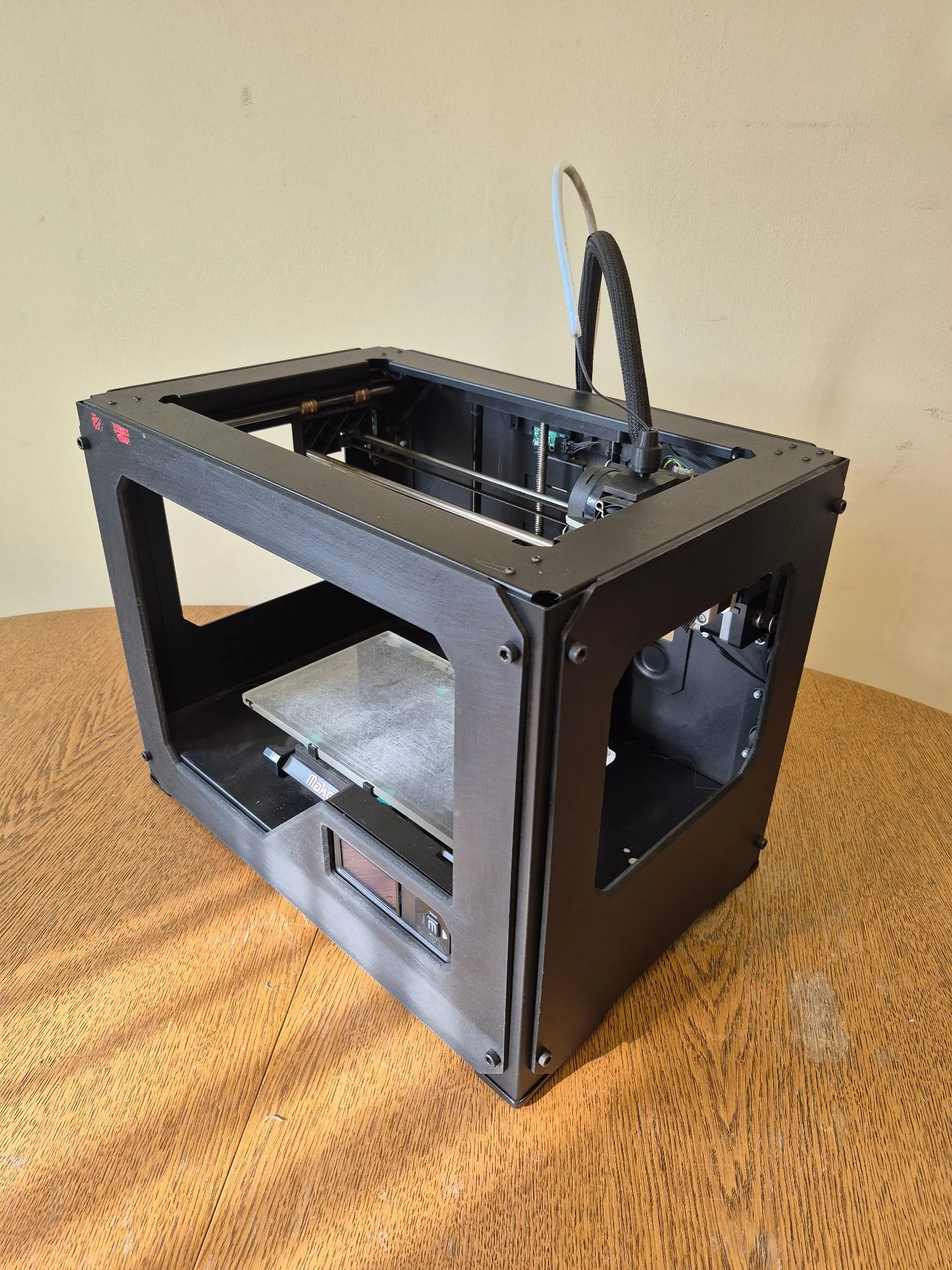 Czarna, sześcianowa obudowa drukarki 3D ustawiona na drewnianym stole. Wewnątrz obudowy widoczny jest stół roboczy oraz mechanizmy drukarki z czarnym kablem prowadzącym do góry. Na przednim panelu znajduje się mały ekran LCD.