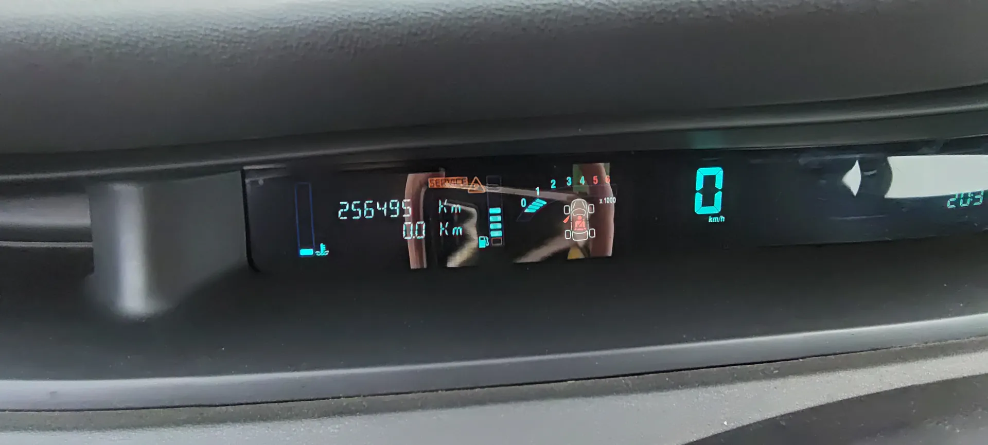 Tablica rozdzielcza samochodu z cyfrowym wyświetlaczem. Po lewej stronie wskaźnik paliwa z niebieskim symbolem, obok licznik przebiegu pokazujący 256495 km. W centralnej części komunikat SERVICE z pomarańczowym trójkątem ostrzegawczym, wskaźnik bieżącego przebiegu 00 km, wskaźnik biegów z oznaczeniami 0, 1, 2, 3, 4, 5, 6 oraz ikoną samochodu z pasem bezpieczeństwa. Po prawej stronie prędkość 0 km/h, temperatura 20,5°C i godzina 20:53. Wyświetlacz ma czarne tło z niebieskimi i pomarańczowymi elementami.