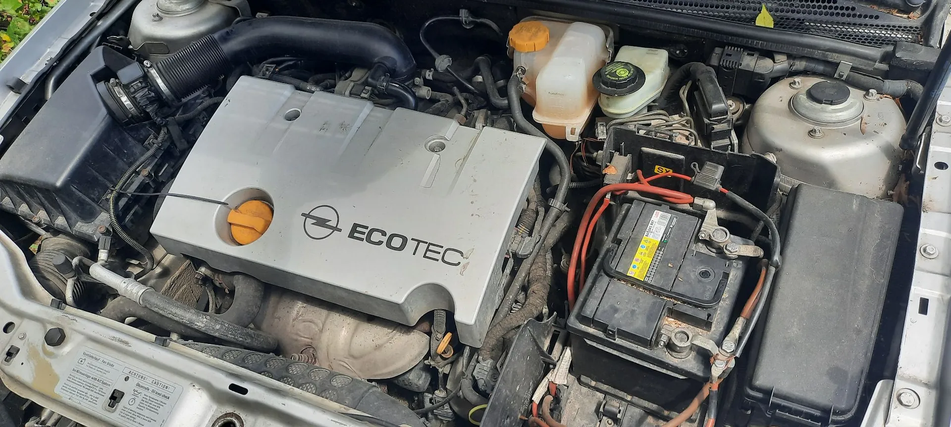 Widok z góry na komorę silnika samochodu. W centrum znajduje się srebrna osłona silnika z logo marki Opel oraz napisem ECOTEC. Po prawej stronie widoczny jest czarny akumulator z żółtą etykietą, obok niego znajduje się zbiornik z płynem spryskiwaczy z żółtym korkiem. Po lewej stronie widoczny jest czarny wlot powietrza i rury doprowadzające. Całość jest osadzona w srebrnej karoserii pojazdu.