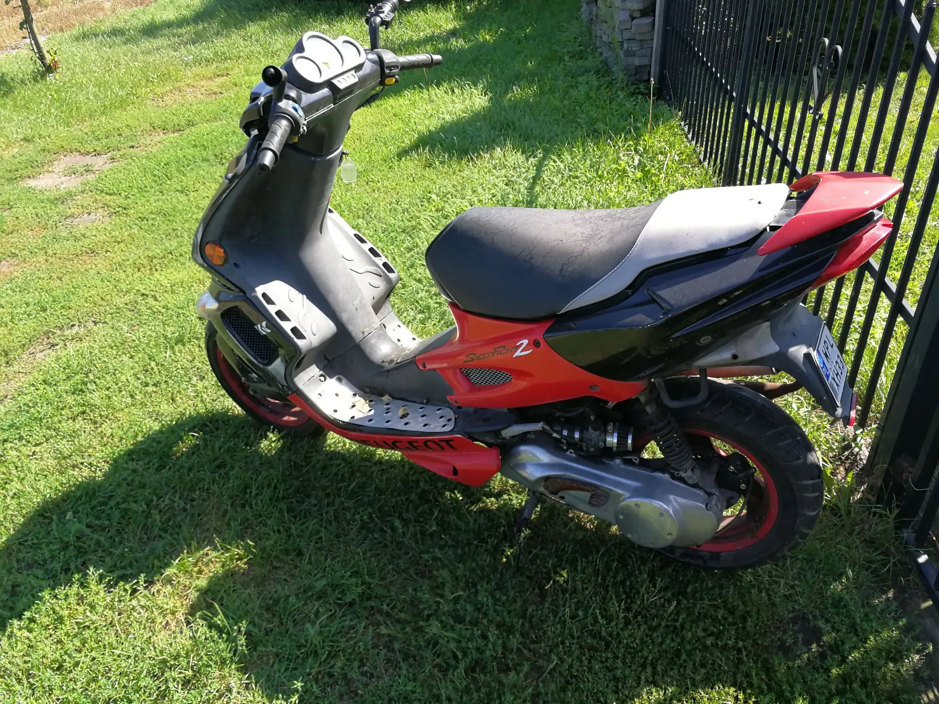 Czerwono-czarny skuter marki Peugeot Speedfight 2 zaparkowany na zielonej trawie obok czarnego metalowego ogrodzenia.