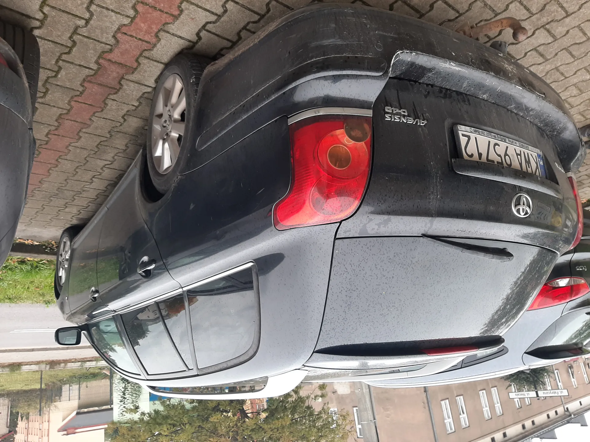 Czarny samochód osobowy typu kombi marki Toyota Avensis D-4D zaparkowany na brukowanej powierzchni. Widok z tyłu, z widocznym tablicą rejestracyjną KWA 95712 oraz logo marki. Samochód pokryty jest kroplami wody. W tle inne zaparkowane pojazdy, budynki mieszkalne, drzewa i znaki uliczne. Nawierzchnia parkingu składa się z czerwono-szarych płytek.