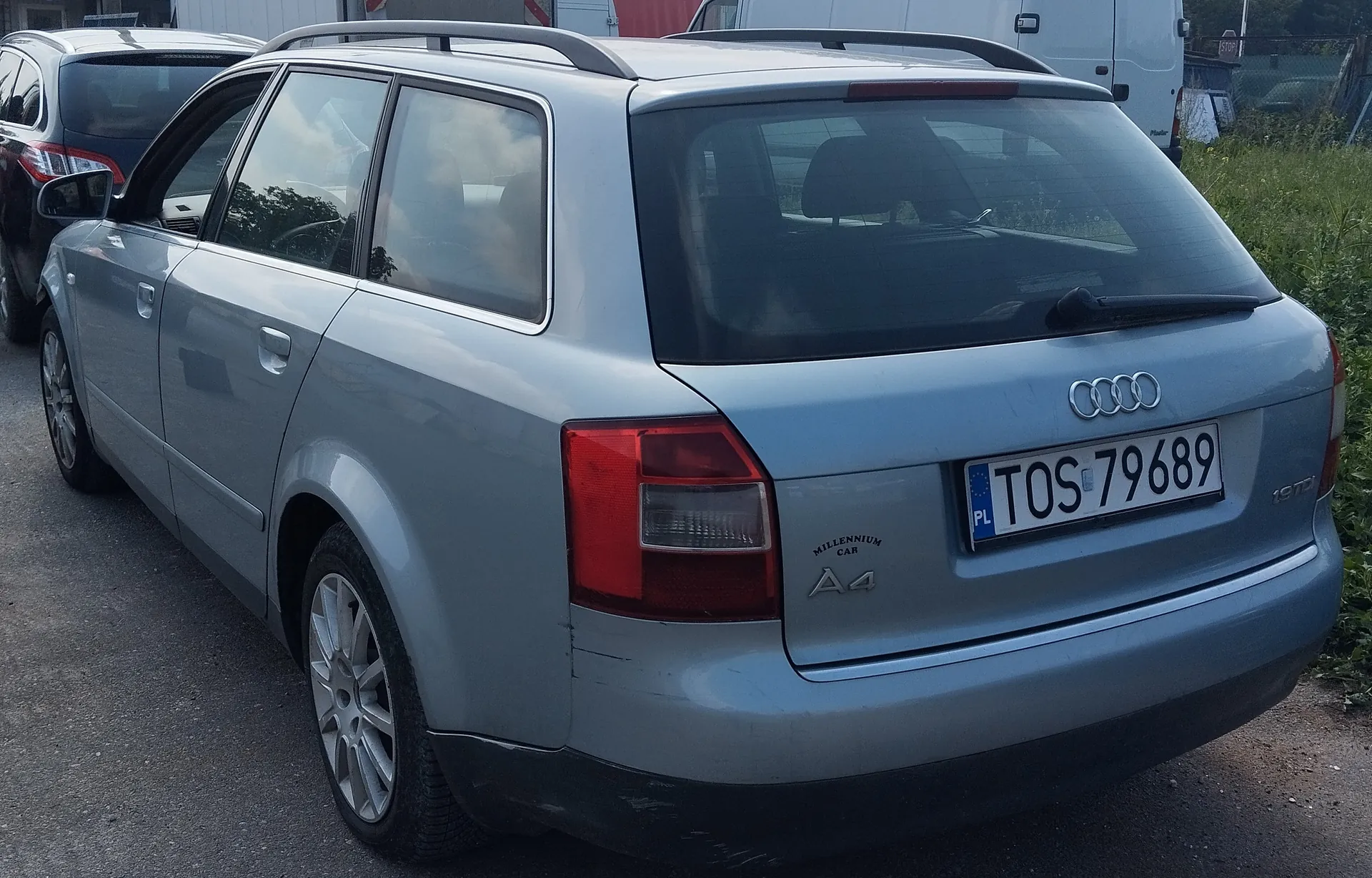 Srebrny samochód Audi A4 kombi z tablicą rejestracyjną PL TOS 79689. Na tylnej części widoczne emblematy Audi, napisy 'Millennium Car' oraz 'A4'. Samochód wyposażony w dachowy bagażnik. W tle inne pojazdy i zielona trawa.